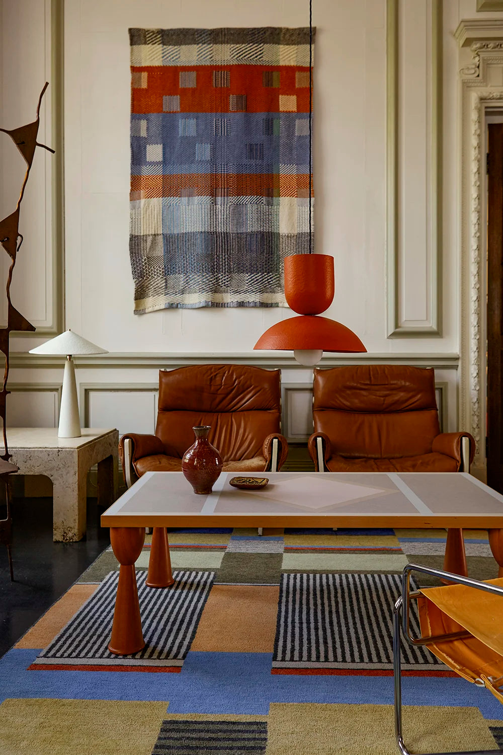 A styled interior featuring two brown leather armchairs, a coffee table, striped rugs, and a blue and orange patterned textile hanging on the wall. A white table lamp sits on a side table with a red-brick pendant hangs above the coffee table.