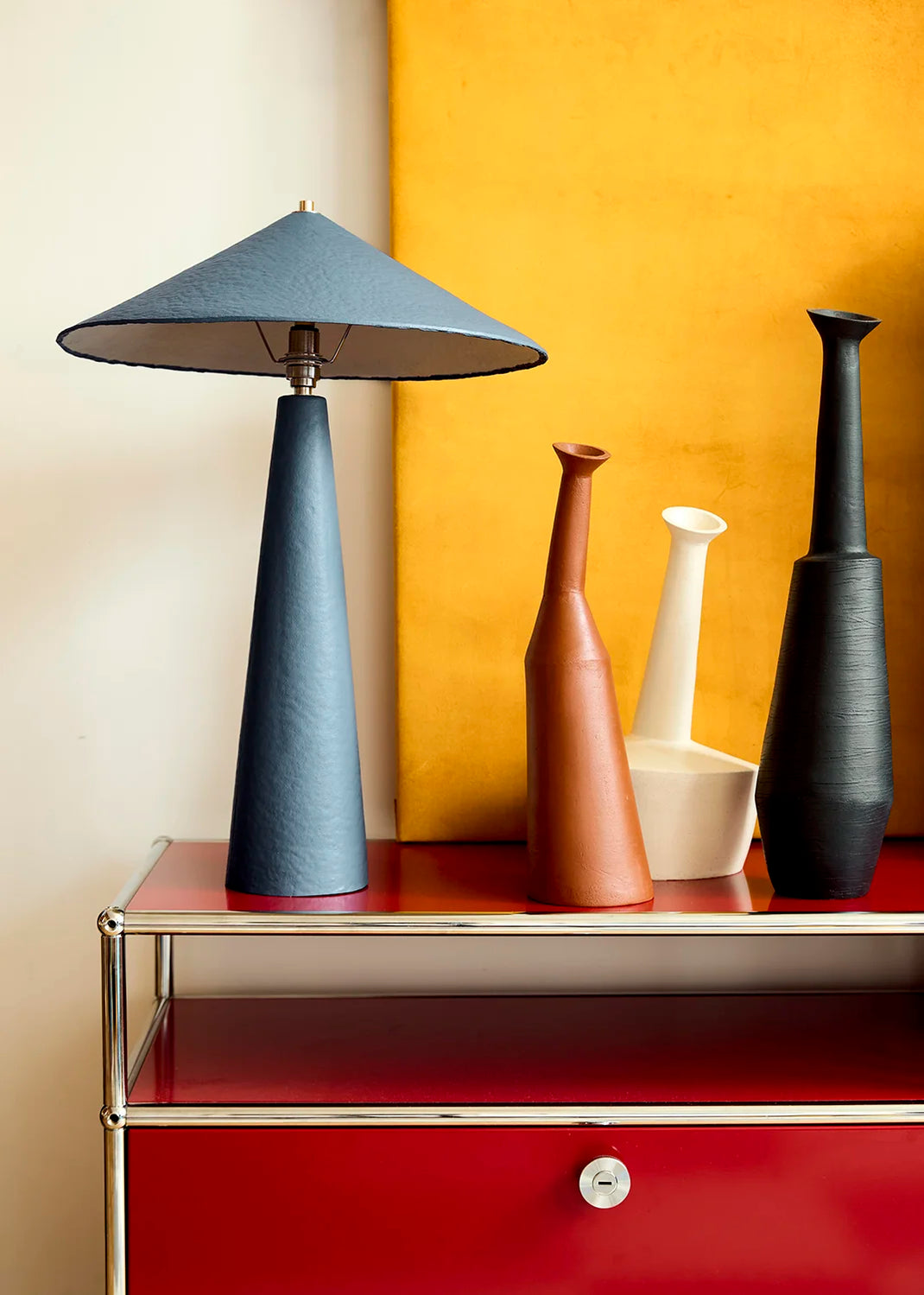 A mid-century modern table lamp with a tapered blue shade and base sits on a red cabinet next to four sculptural ceramic vases in terracotta, white, and black against a split cream and ochre background.