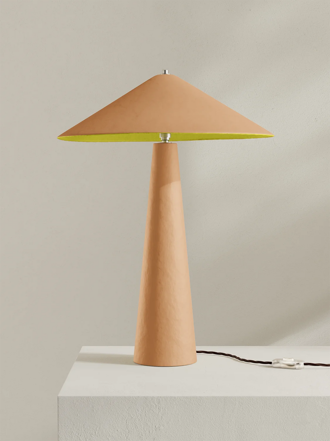 A modern table lamp with a tapered peach-colored shade and base, featuring a bright yellow inner shade, shown on a white pedestal against a neutral beige background.