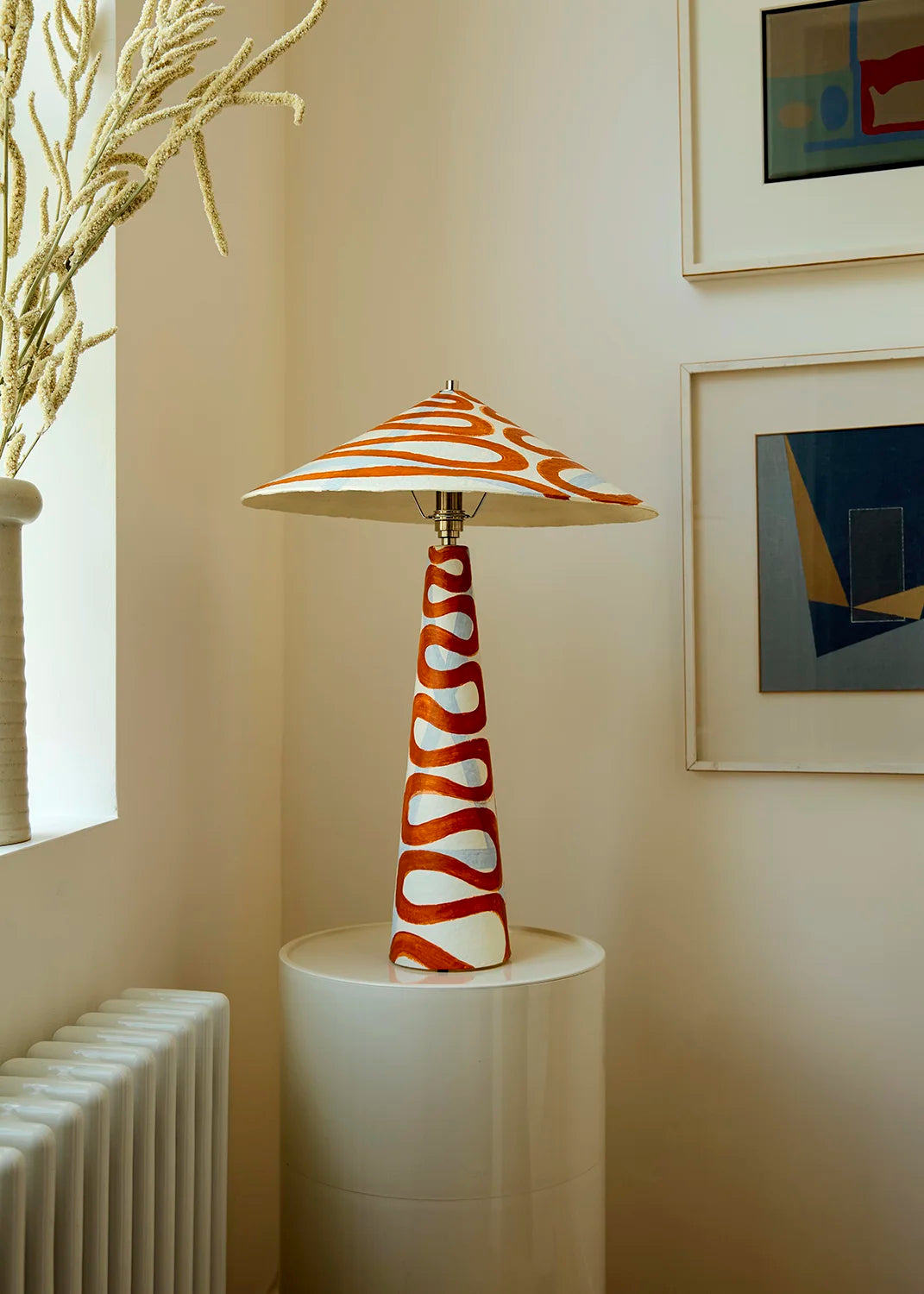 A table lamp with a tapered white shade featuring orange wavy stripes and a matching orange-and-white patterned base, displayed on a white pedestal in a warm interior setting.