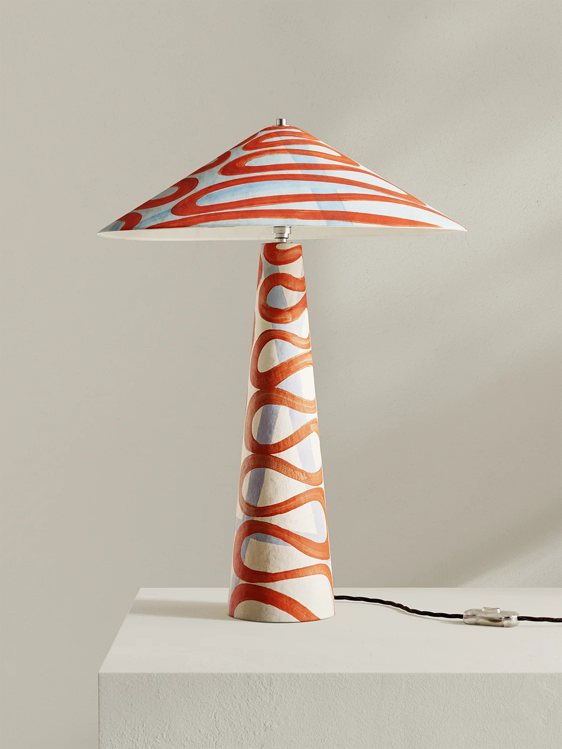 A table lamp with a tapered conical base and a wide conical shade, both decorated with a wavy, ribbon-like pattern in terracotta orange, light blue, and white. The lamp is displayed on a white pedestal against a neutral beige background.