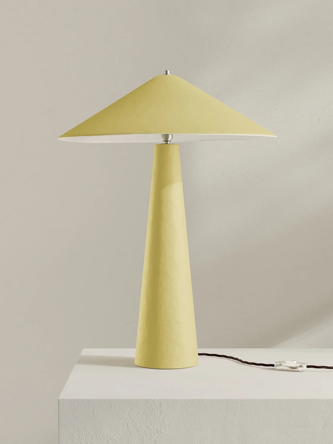 A modern table lamp with a tapered yellow shade and matching yellow base, shown on a white pedestal against a light beige wall.