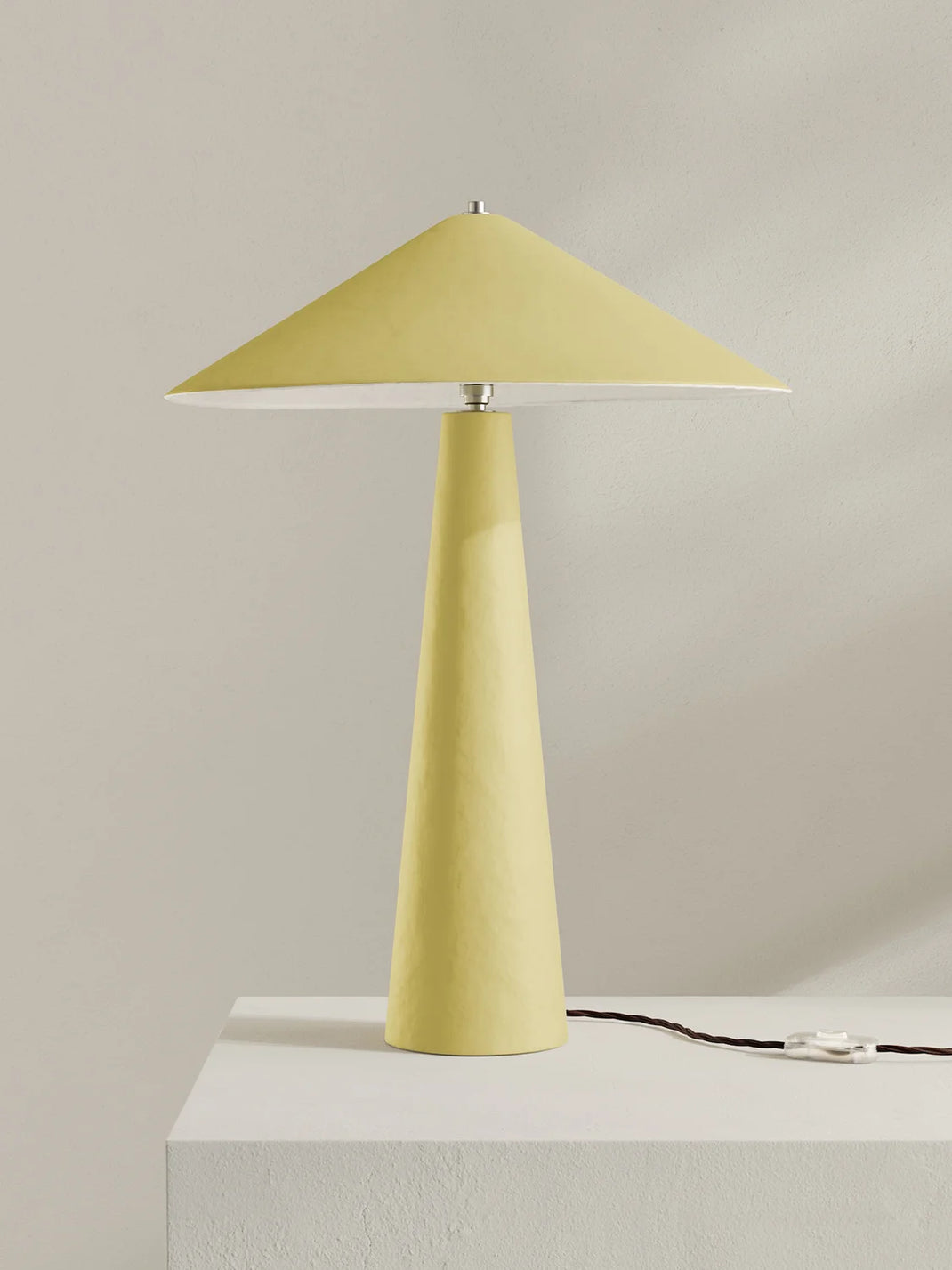 A modern table lamp with a tapered yellow shade and matching yellow base, shown on a white pedestal against a light beige wall.
