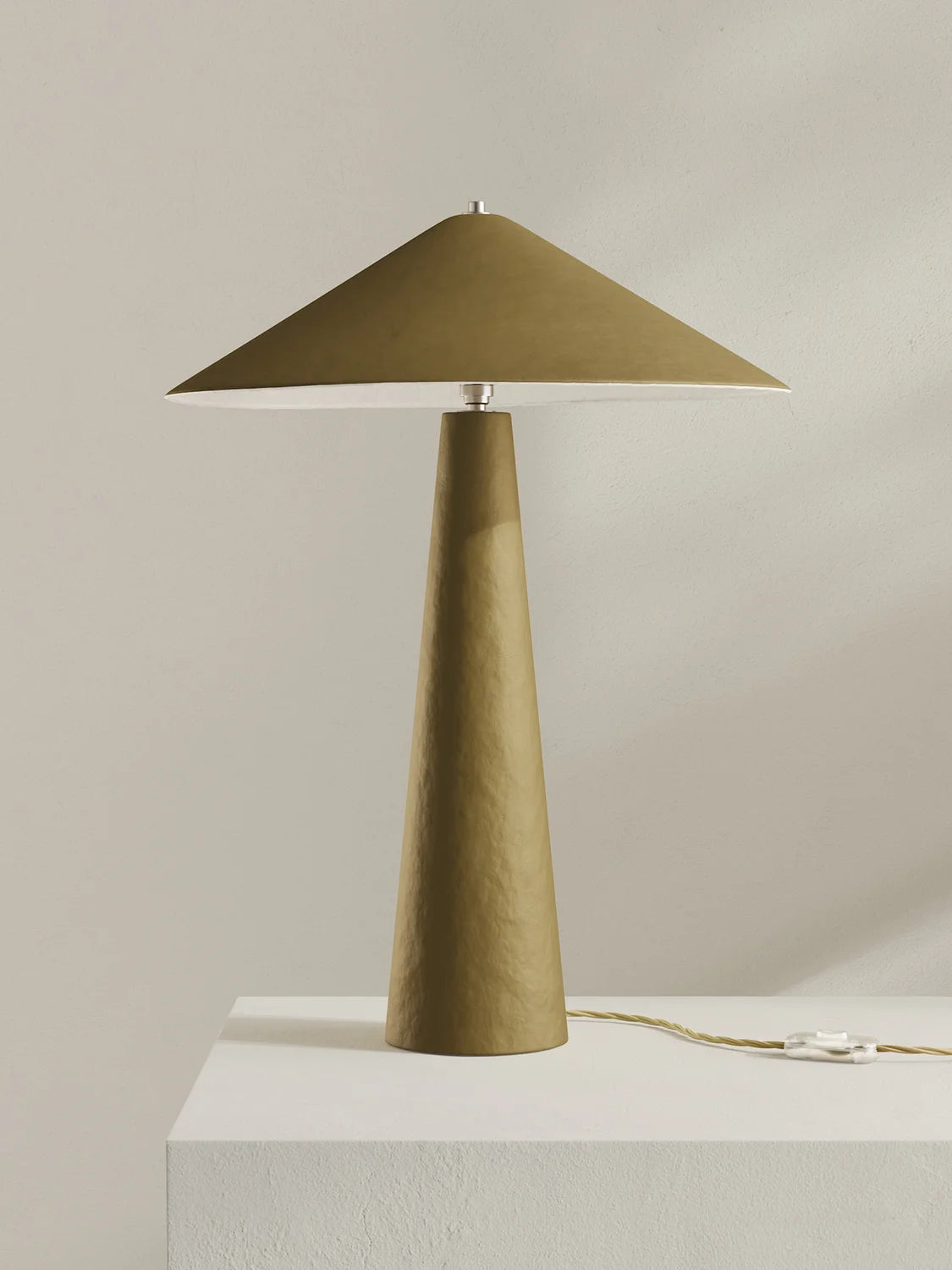 A modern table lamp with a tapered moss-green base and a matching wide-brimmed shade, displayed on a white pedestal against a neutral beige background.