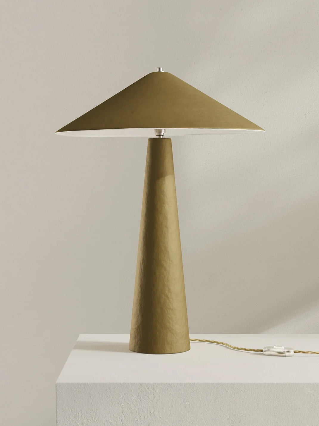 A modern table lamp with a tapered moss-green base and a matching wide-brimmed shade, displayed on a white pedestal against a neutral beige background.