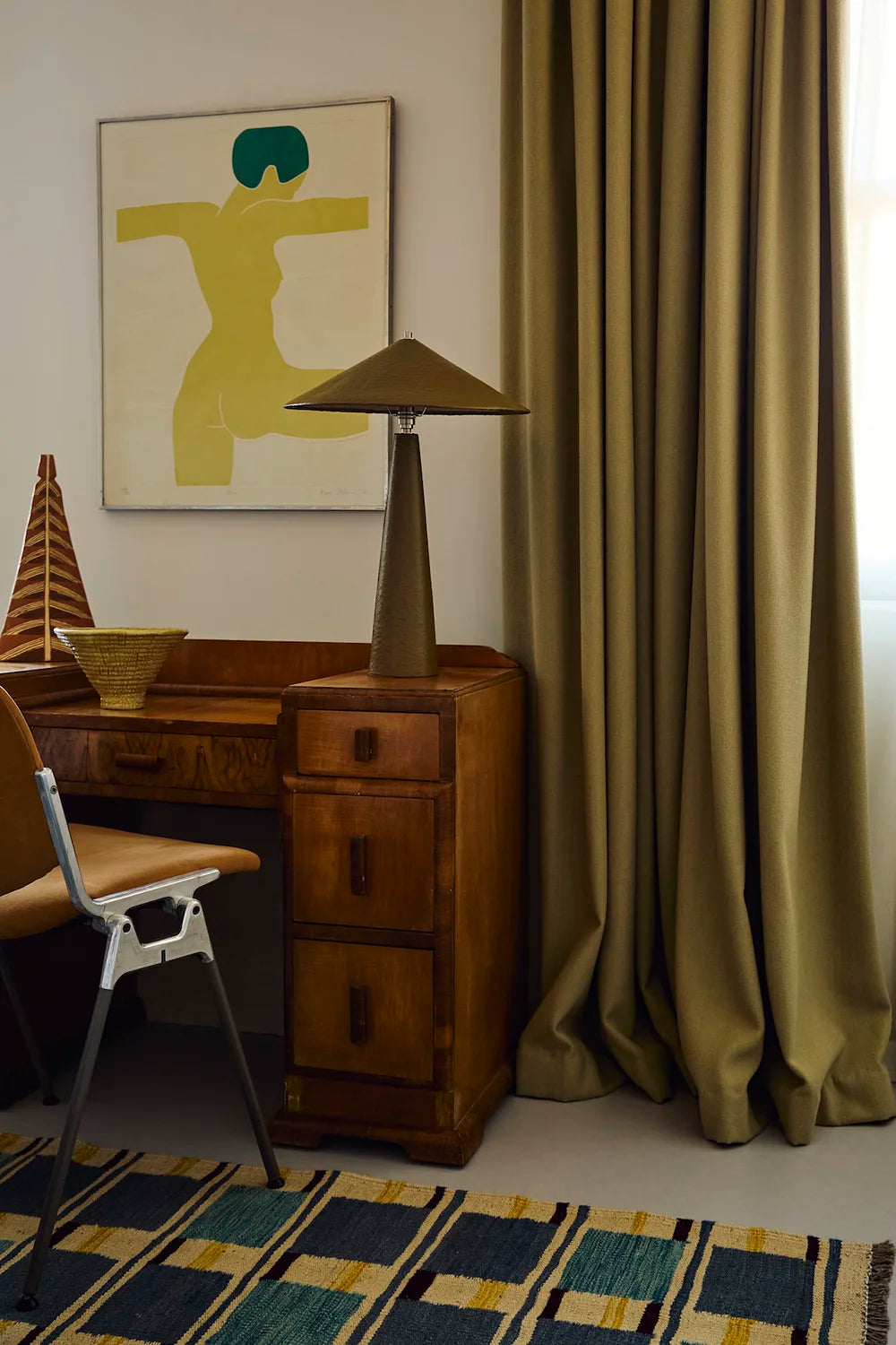 A styled interior corner featuring a pair of floor-length olive green curtains next to a wooden desk, moss-green table lamp, chair, and a geometric patterned rug.