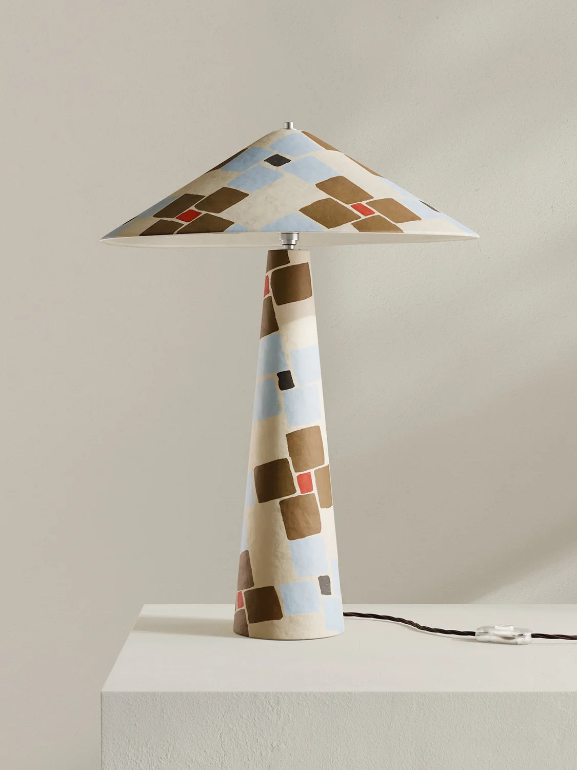 A table lamp with a tapered conical base and matching wide shade, decorated with an abstract geometric pattern of brown, beige, light blue, and small red squares, shown on a white pedestal against a neutral beige background.