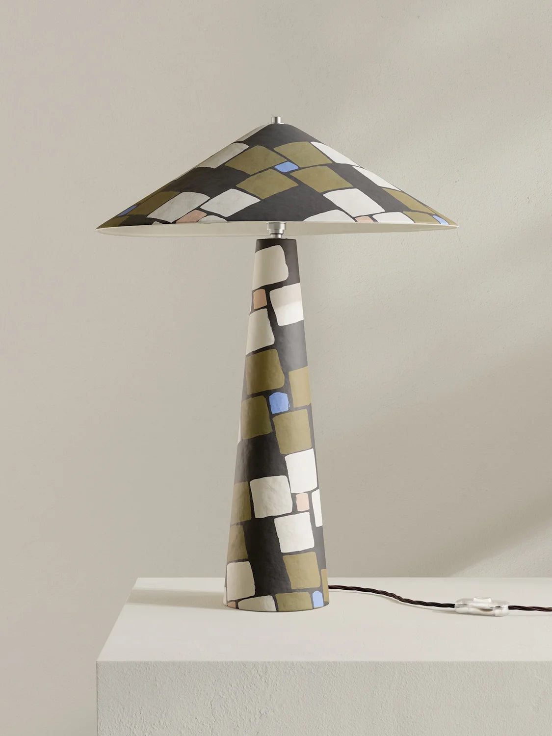A modern table lamp with a tapered conical shade and matching base, featuring an abstract geometric pattern of black, olive green, white, and beige blocks with a small light blue accent. The lamp is shown on a white pedestal against a neutral beige background.