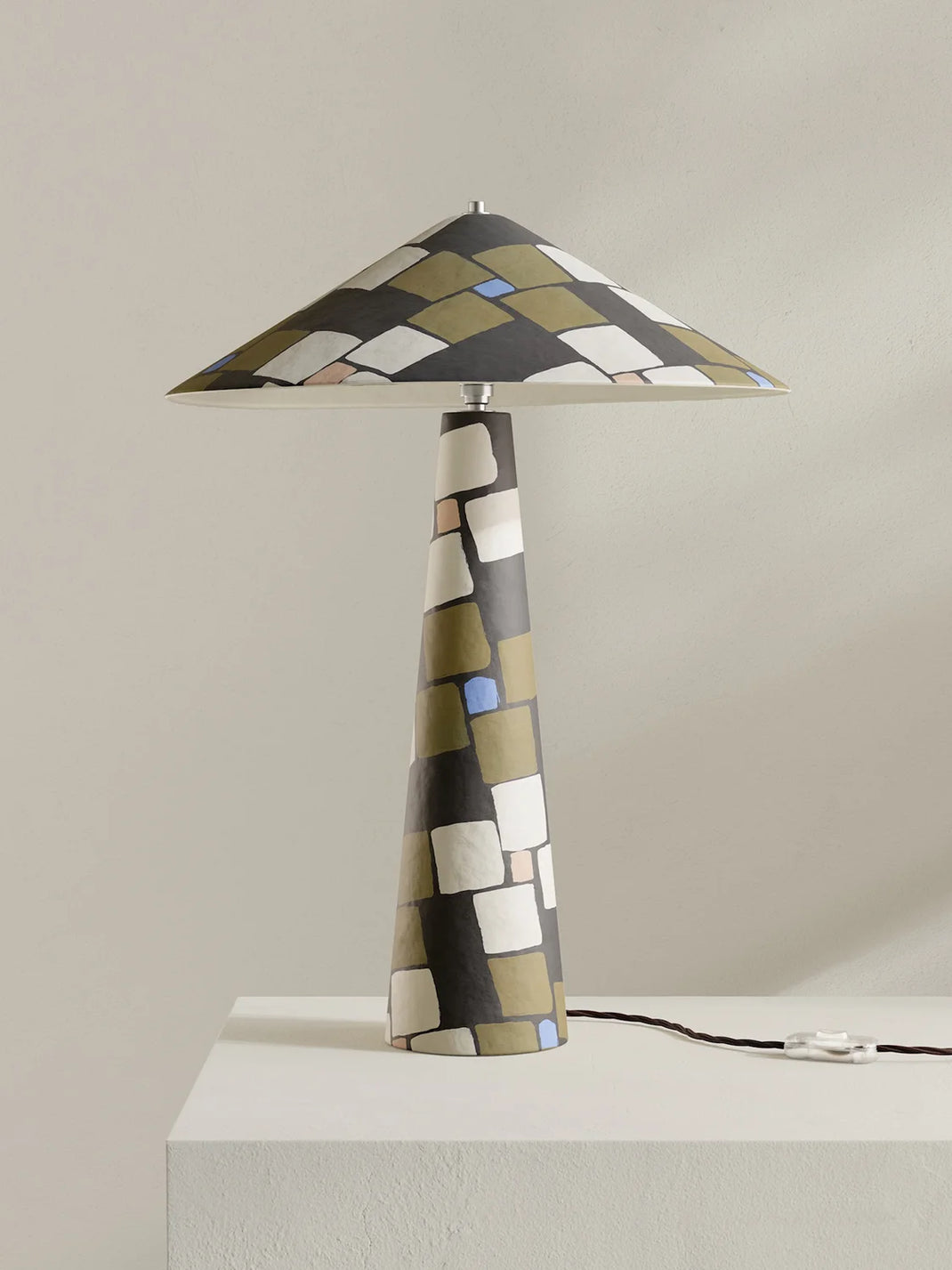 A modern table lamp with a tapered conical shade and matching base, featuring an abstract geometric pattern of black, olive green, white, and beige blocks with a small light blue accent. The lamp is shown on a white pedestal against a neutral beige background.
