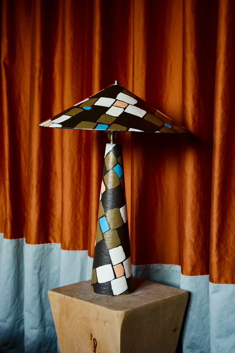 A handmade table lamp with a geometric mosaic pattern in brown, gold, white, and blue, displayed on a wooden block in front of a two-toned orange and blue curtain.