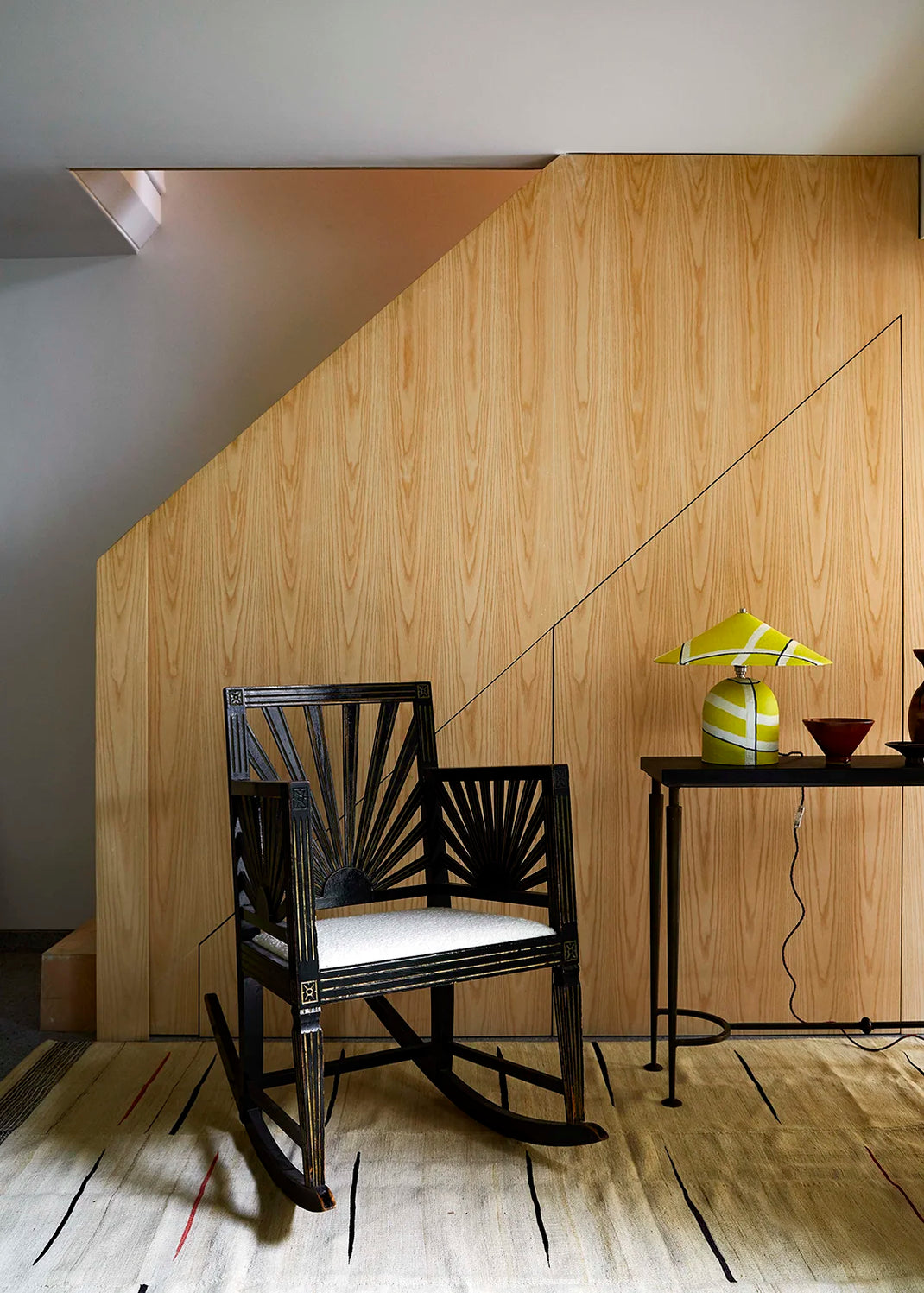 A black rattan rocking chair with a sunburst backrest and white cushion, positioned in front of a light wood-paneled wall in a room setting. A yellow table lamp with a white and navy striped pattern sits on a metal table.