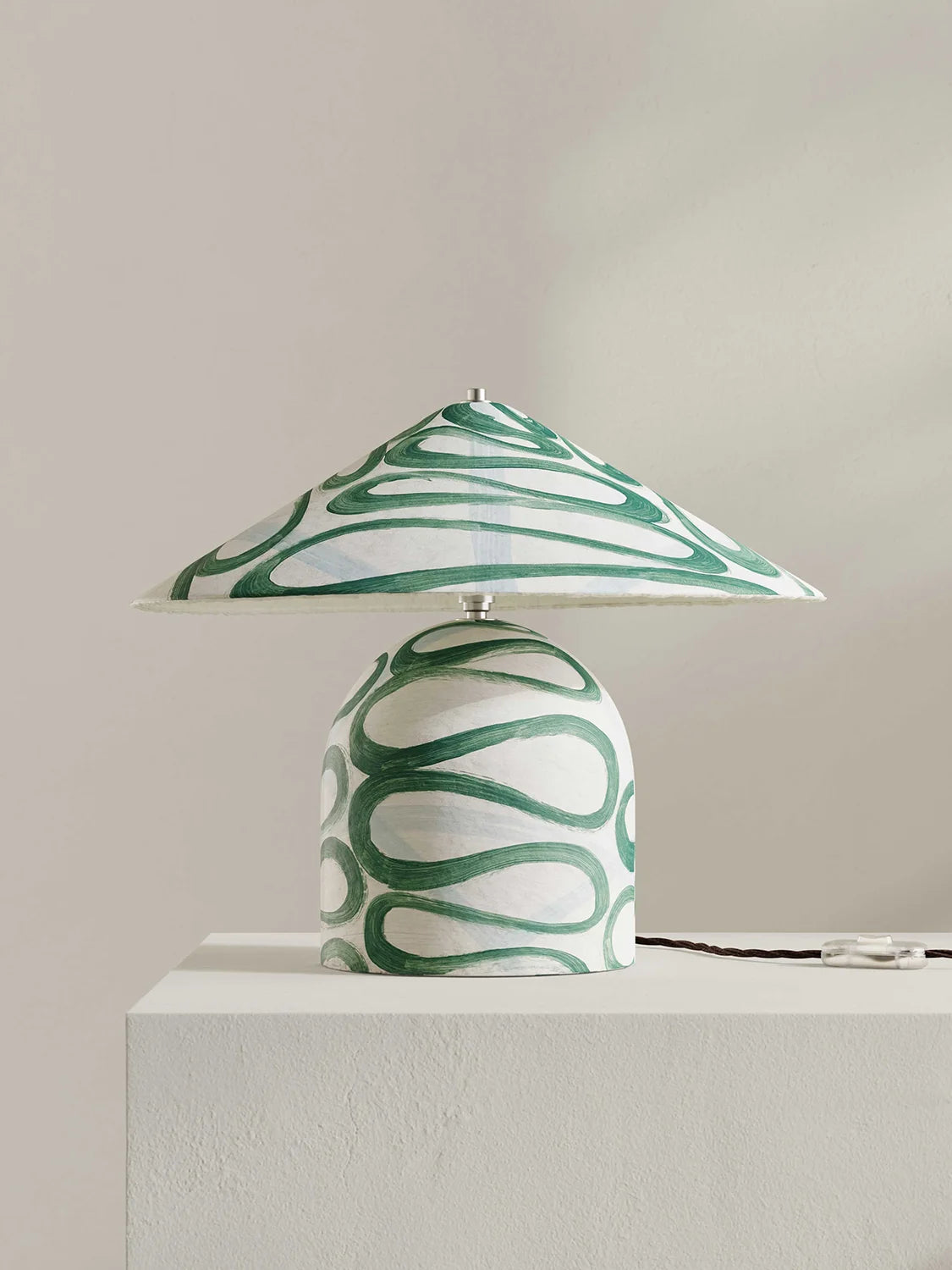 A table lamp with a wide, shallow conical shade and a rounded base, both decorated with a green and white wavy abstract pattern, displayed on a white pedestal against a neutral beige background.