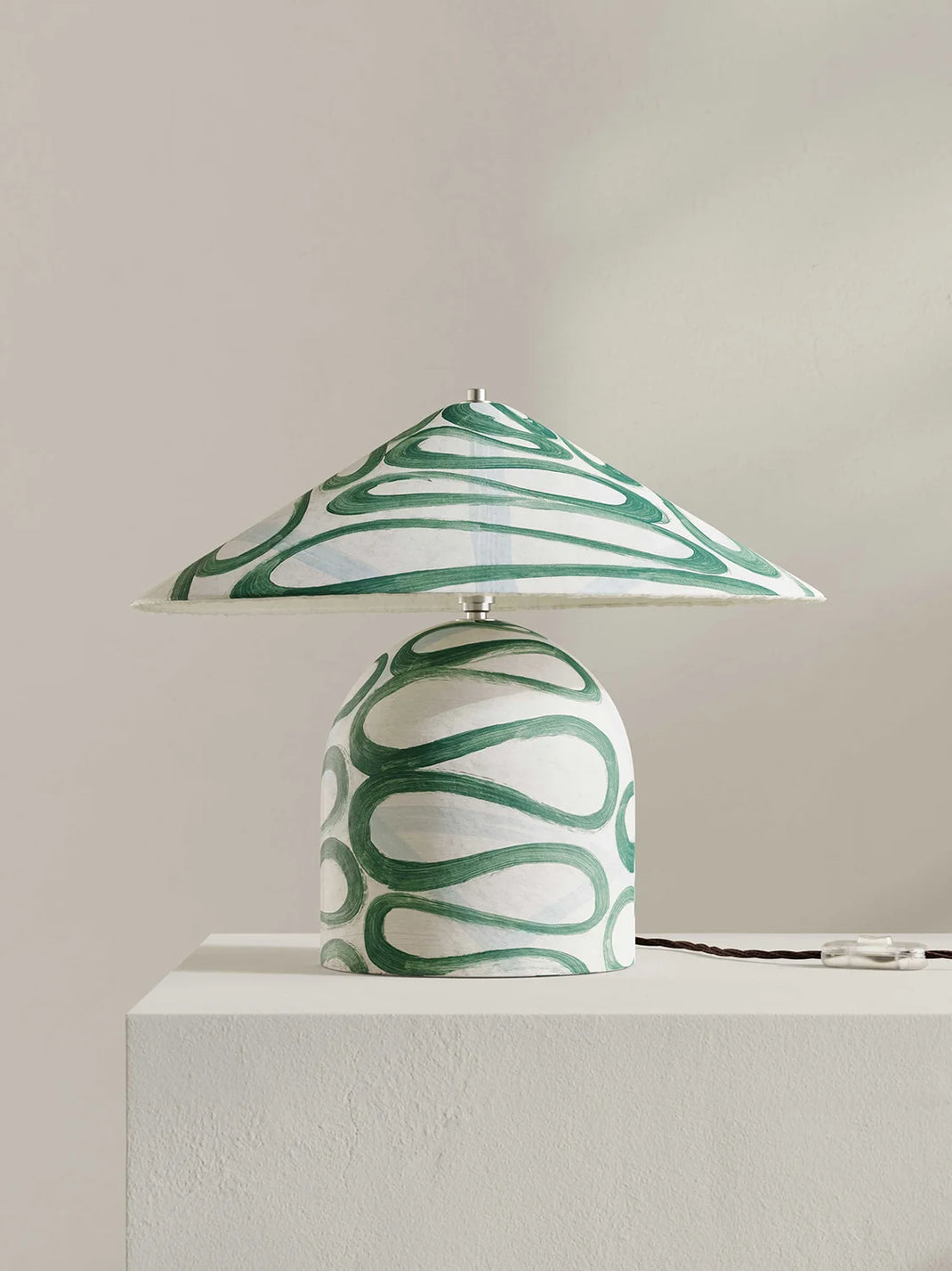 A table lamp with a wide, shallow conical shade and a rounded base, both decorated with a green and white wavy abstract pattern, displayed on a white pedestal against a neutral beige background.