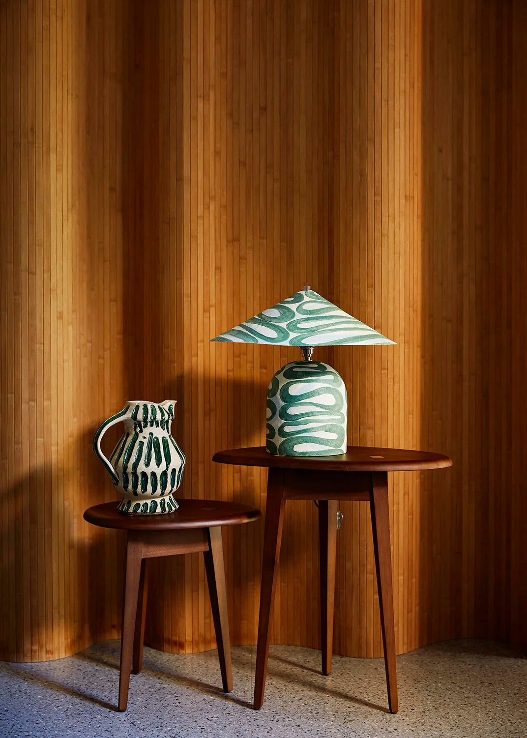 A mid-century modern ceramic table lamp with a green and white wavy patterned shade and base sits on a small round wooden side table, next to a matching ceramic pitcher on a matching wooden stool, set against a warm wood-paneled wall.