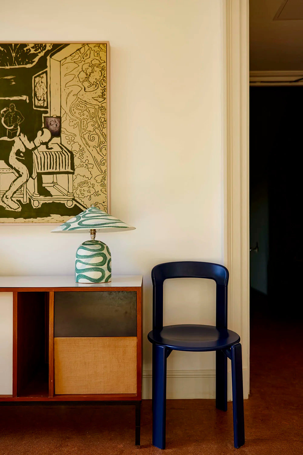 Blue chair next to a wooden cabinet with a lamp and artwork on the wall.