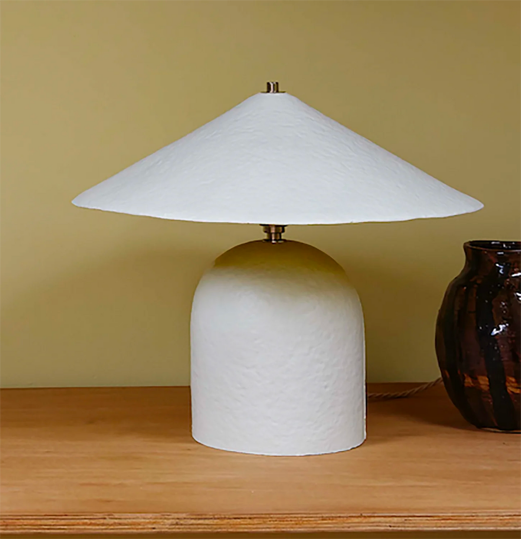 A modern table lamp with a textured white dome-shaped base and a matching wide-brimmed white shade, set on a wooden surface against a warm beige wall.