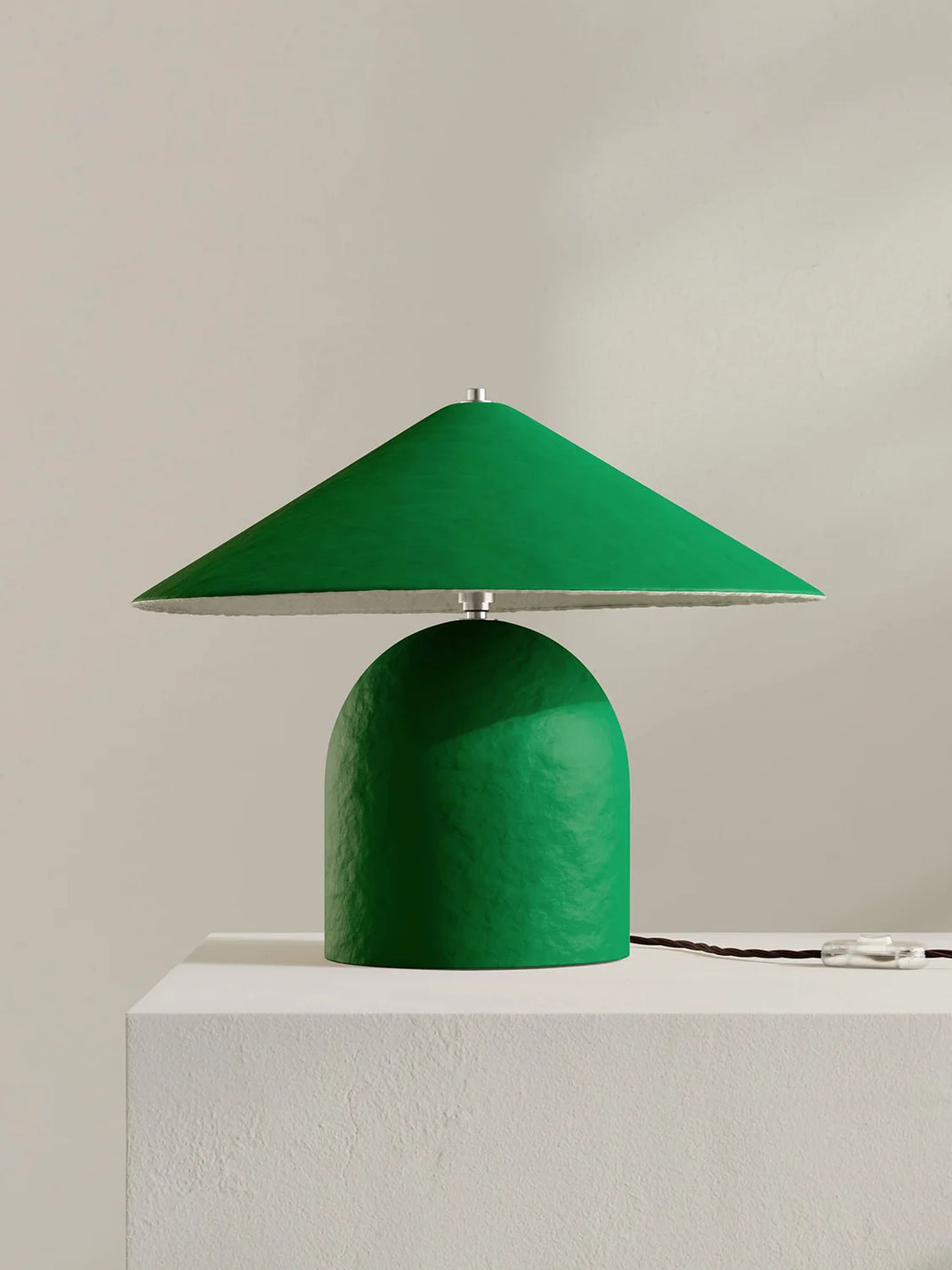 A modern table lamp with a textured green dome-shaped base and a matching green conical shade, shown on a white pedestal against a neutral beige wall.