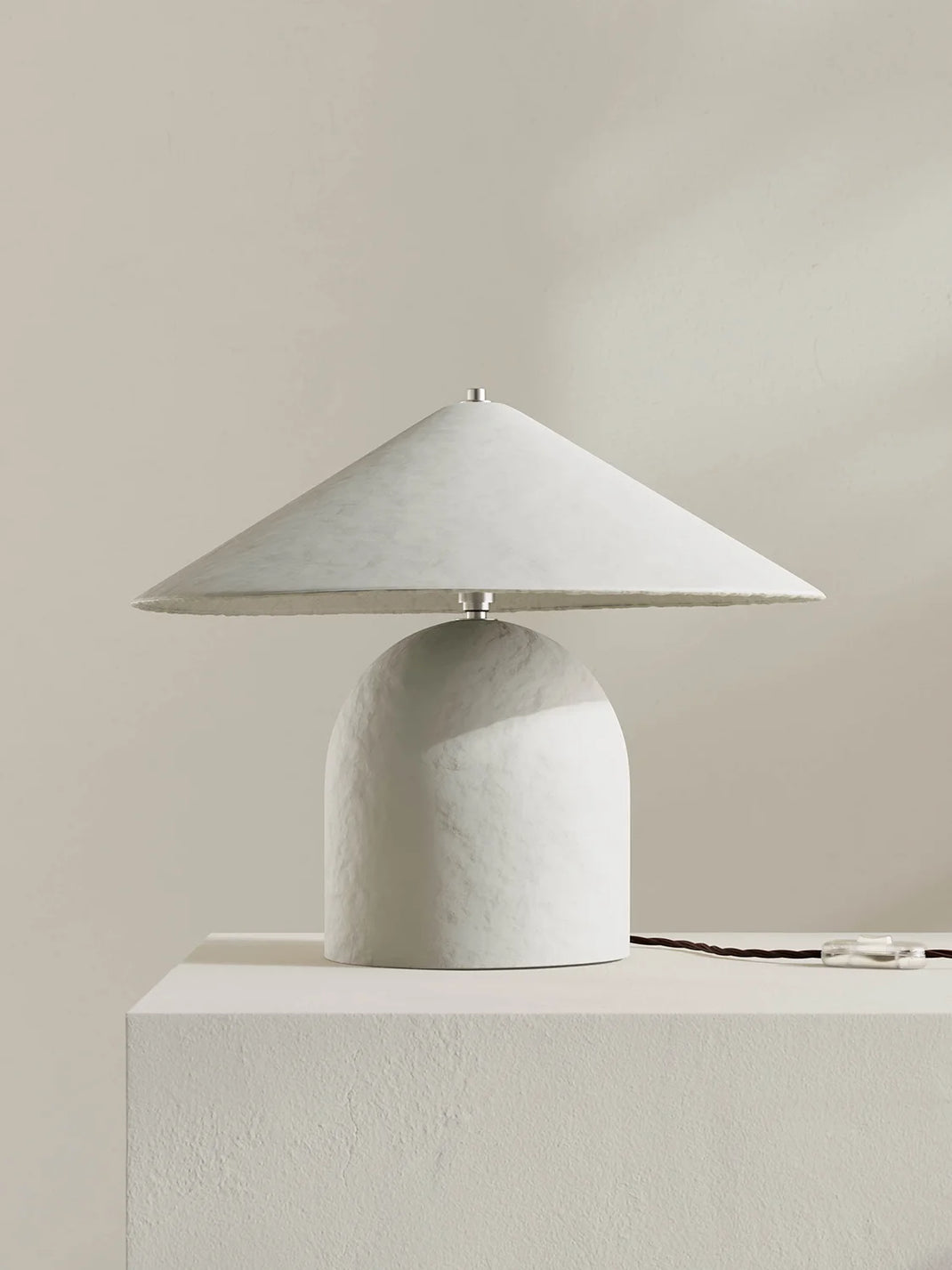A modern table lamp with a wide, shallow conical shade and a rounded textured base, finished in a white color, displayed on a matching pedestal against a neutral beige background.
