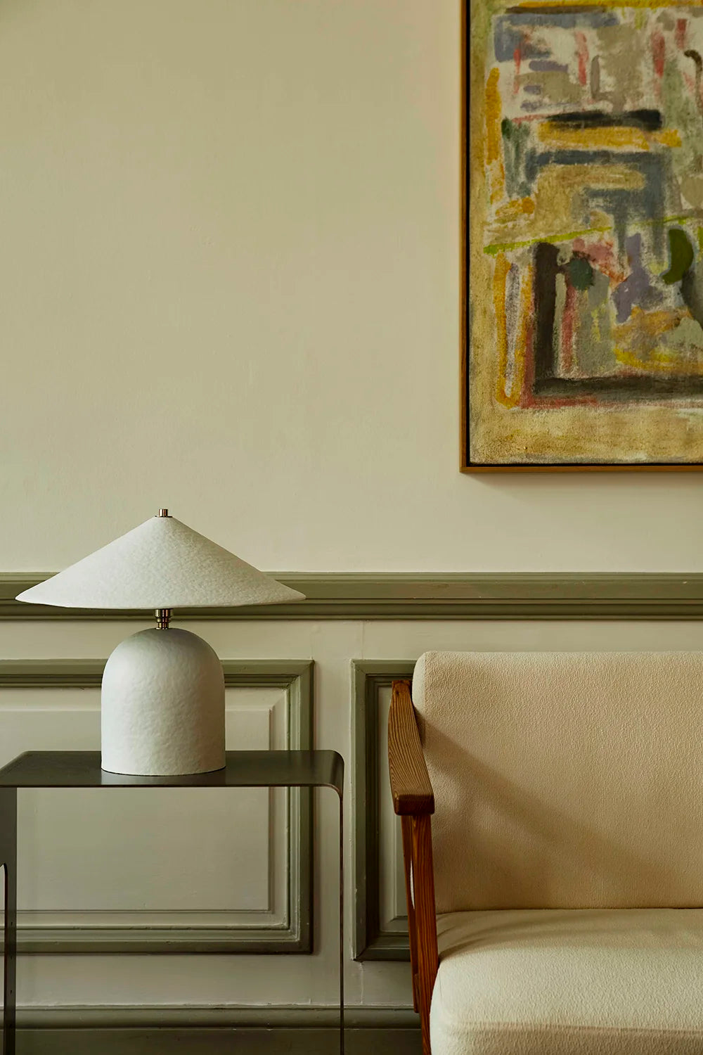 White lamp on a side table next to a beige armchair with abstract art on the wall.