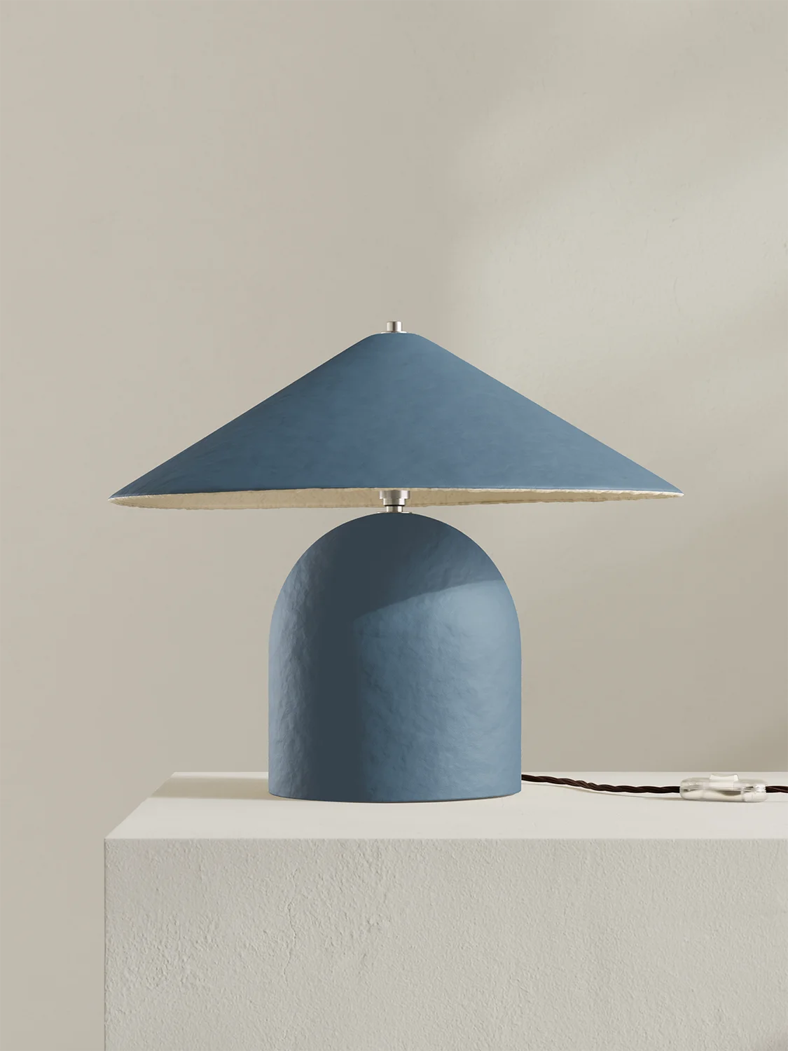 A modern table lamp with a textured blue dome-shaped base and a matching blue conical shade, shown on a white pedestal against a warm beige background.