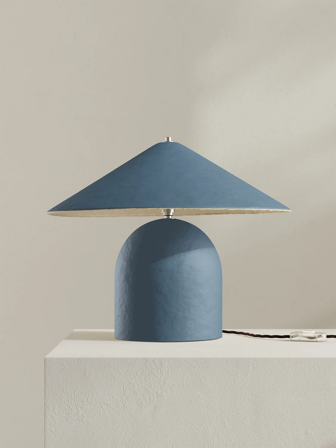 A modern table lamp with a textured blue dome-shaped base and a matching blue conical shade, shown on a white pedestal against a warm beige background.