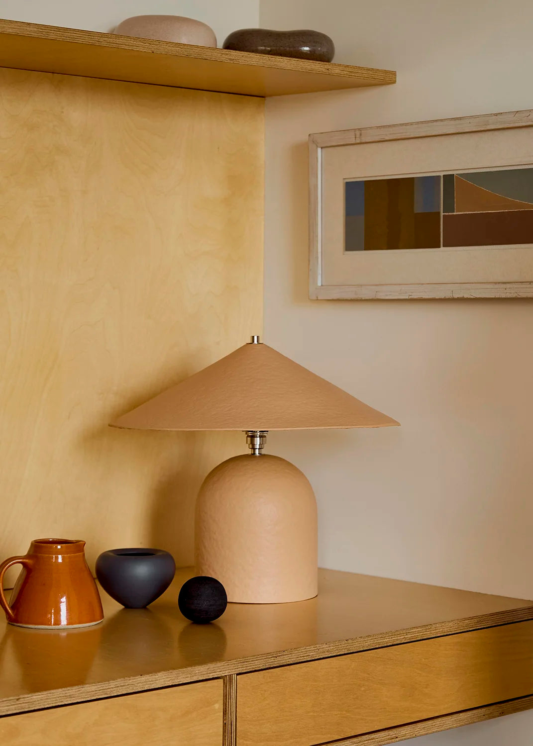Peach coloured table lamp on a wooden surface with decorative items and a framed abstract painting in the background.