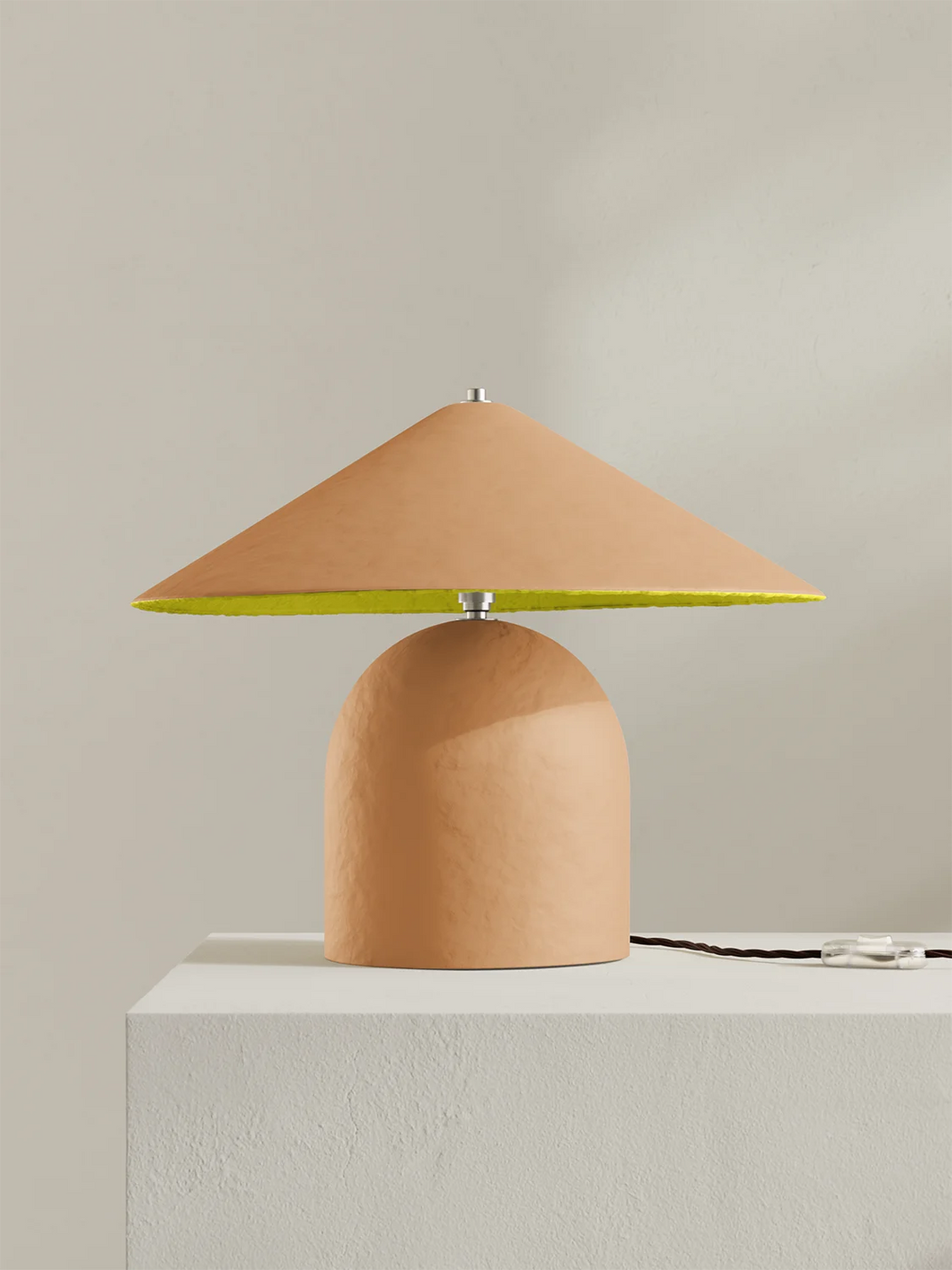 A modern table lamp with a peach-colored dome-shaped base and a matching conical shade, featuring a bright yellow underside on the shade and a silver metal finial, displayed on a white pedestal against a neutral beige background.