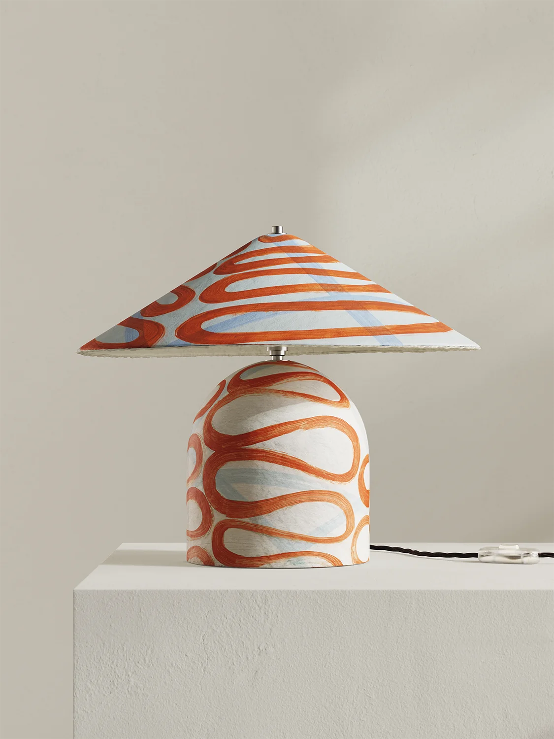 A table lamp with a conical shade and rounded base, both decorated with an orange and white wavy pattern, displayed on a white pedestal against a neutral beige background.