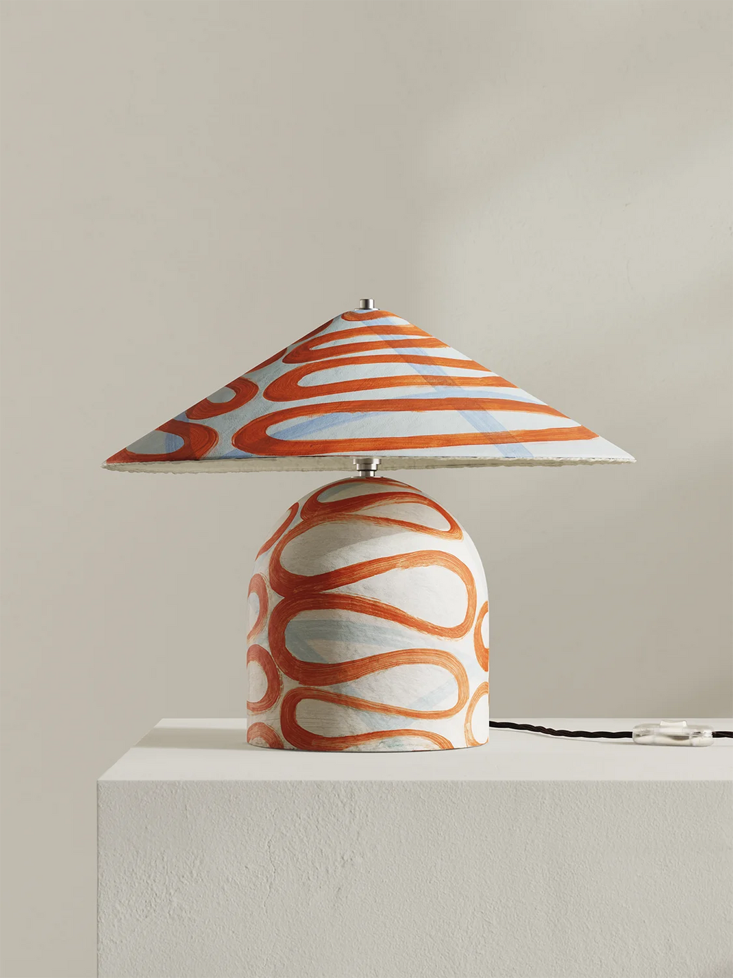 A table lamp with a conical shade and rounded base, both decorated with an orange and white wavy pattern, displayed on a white pedestal against a neutral beige background.