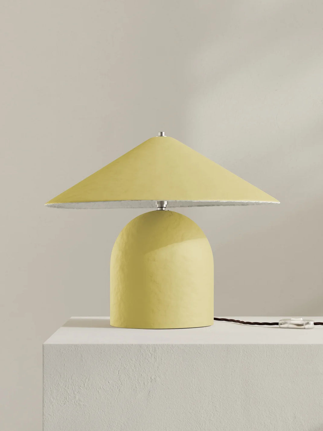 A modern table lamp with a muted yellow dome-shaped base and a matching wide-brimmed cone shade, shown on a white pedestal against a warm beige wall.