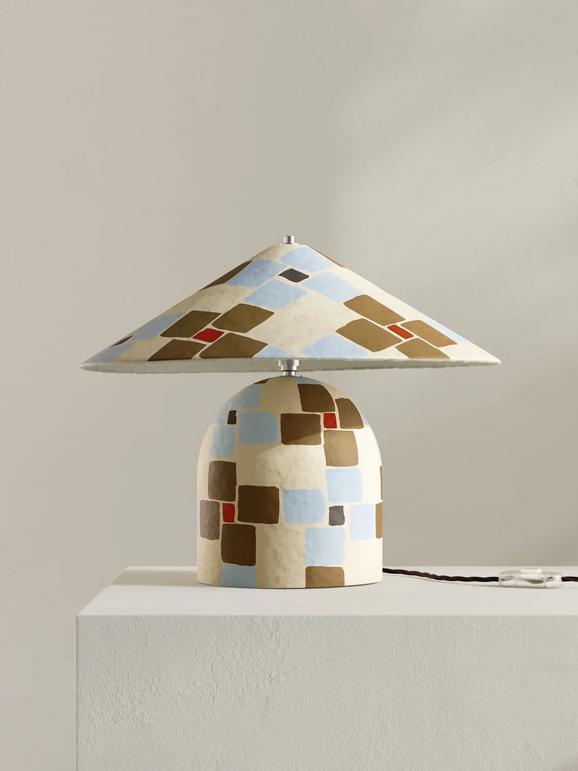 A table lamp with a wide, shallow conical shade and a rounded base, both decorated with a geometric patchwork pattern of beige, brown, light blue, and small red squares. The lamp is displayed on a white pedestal against a neutral beige background.
