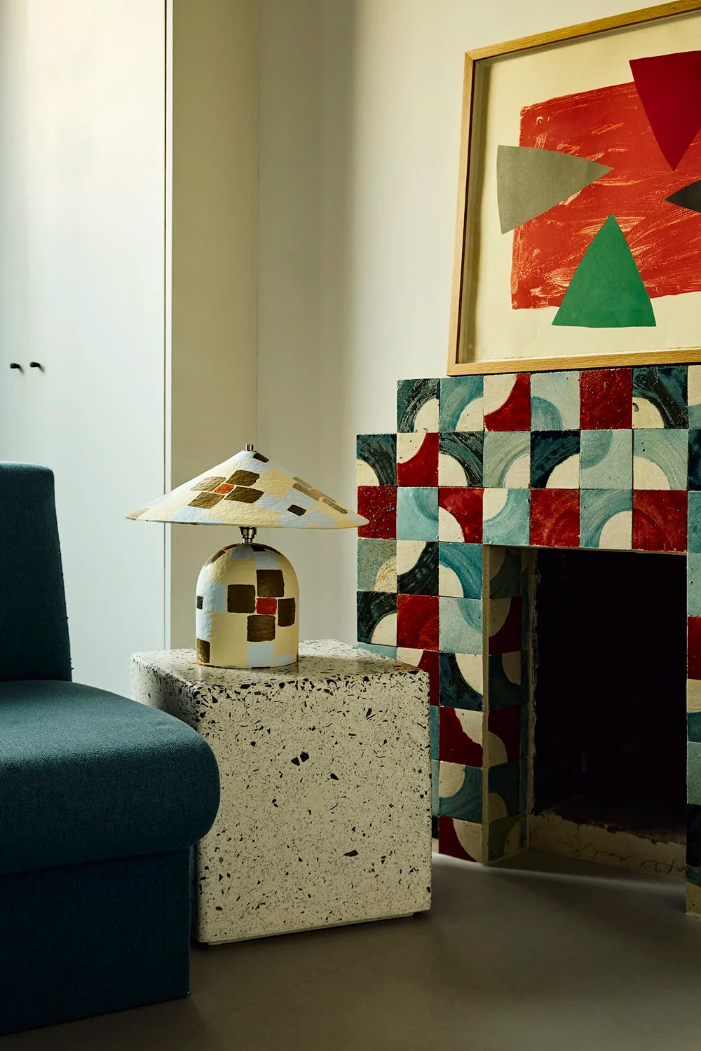 A modern interior scene featuring a terrazzo side table with a small patterned ceramic table lamp, next to a fireplace with a geometric patterned tile surround and a large abstract art print hanging above.