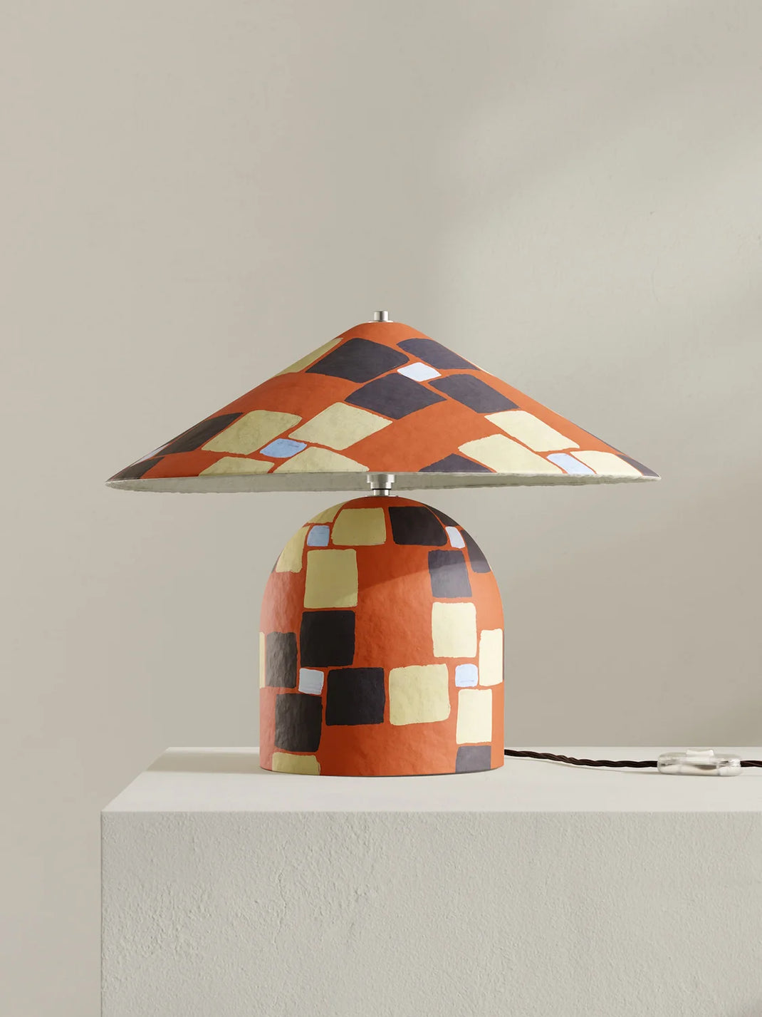 A table lamp with a wide conical shade and a rounded base, both decorated with a geometric mosaic pattern of orange, black, beige, and white squares, displayed on a white pedestal against a neutral beige background.