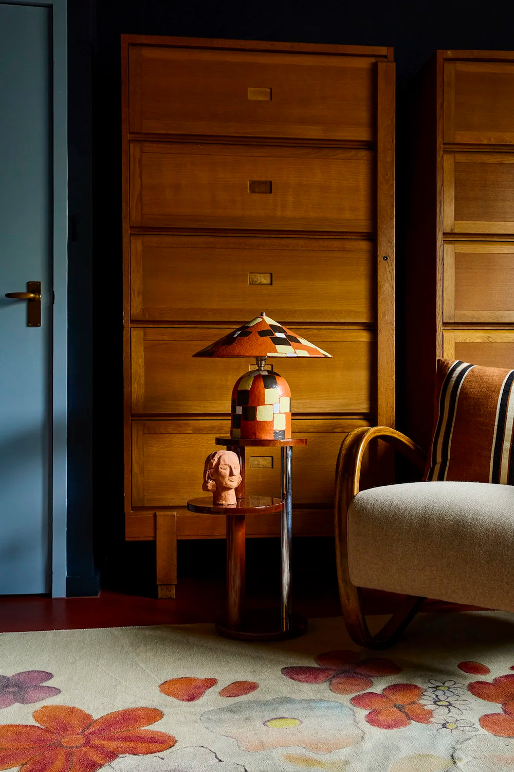 A mid-century modern scene featuring a tall wooden chest of drawers, a small side table with a colorful mosaic table lamp and a small figurine, and part of a striped armchair on a floral rug.