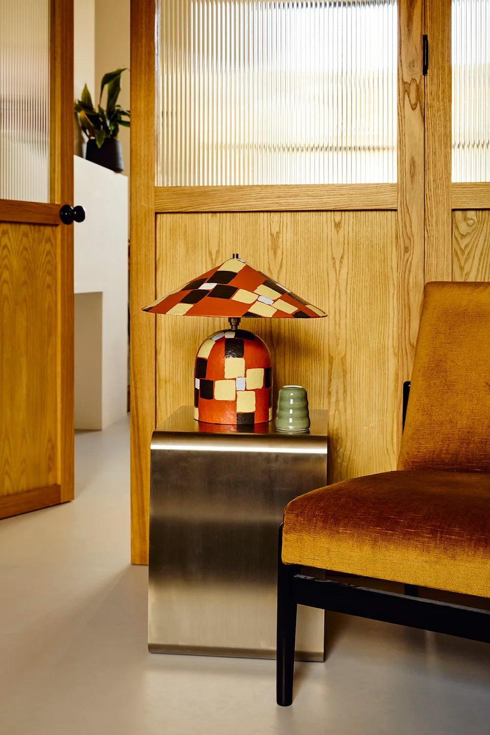 A mid-century modern table lamp with a geometric patchwork shade and base in orange, black, and cream, styled on a brushed metal side table next to a mustard yellow armchair in a warm wood-paneled room.
