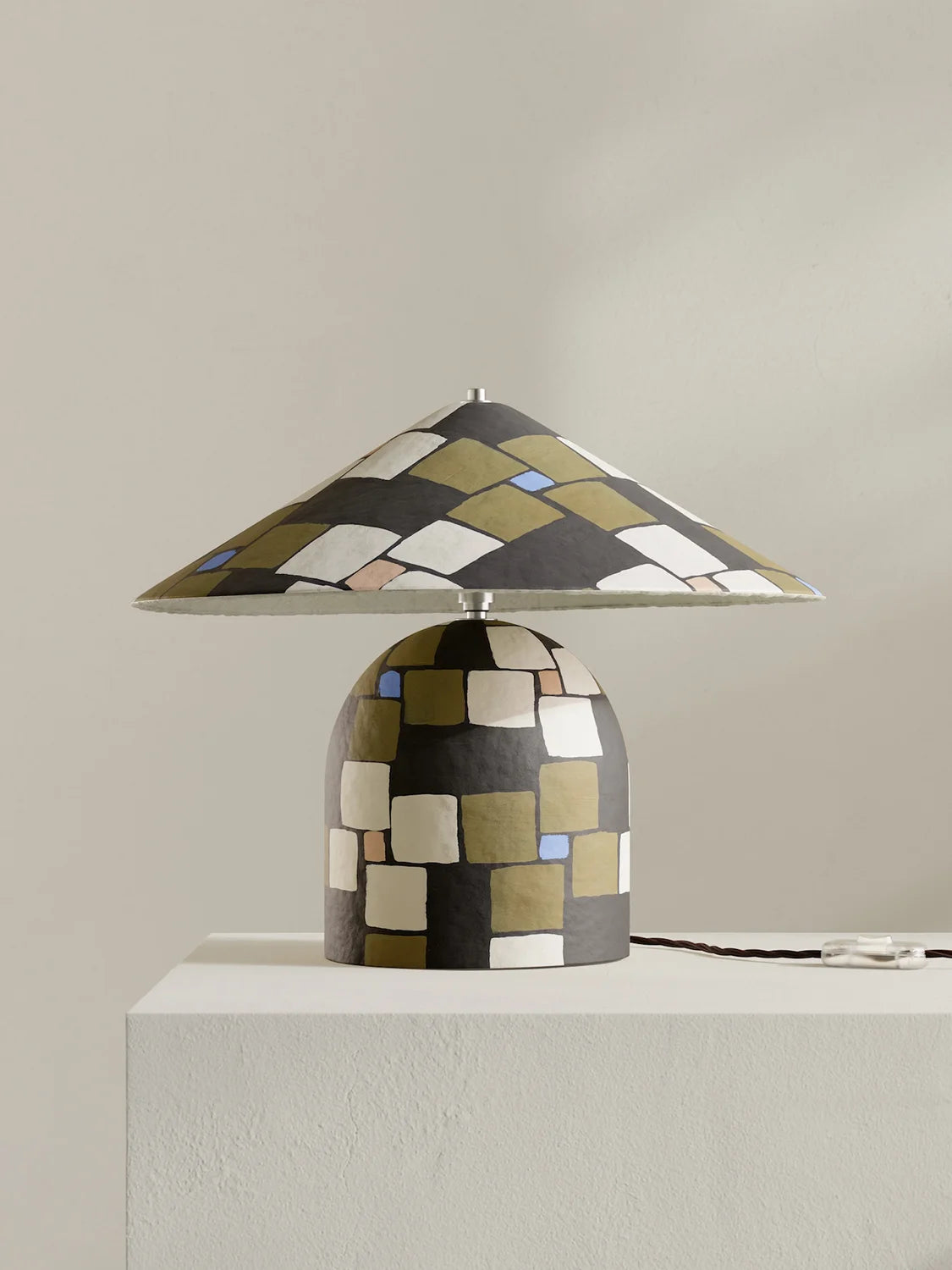 A table lamp with a wide conical shade and a rounded base, both decorated with a geometric mosaic pattern in black, olive green, beige, and small blue accents, displayed on a white pedestal against a neutral beige wall.