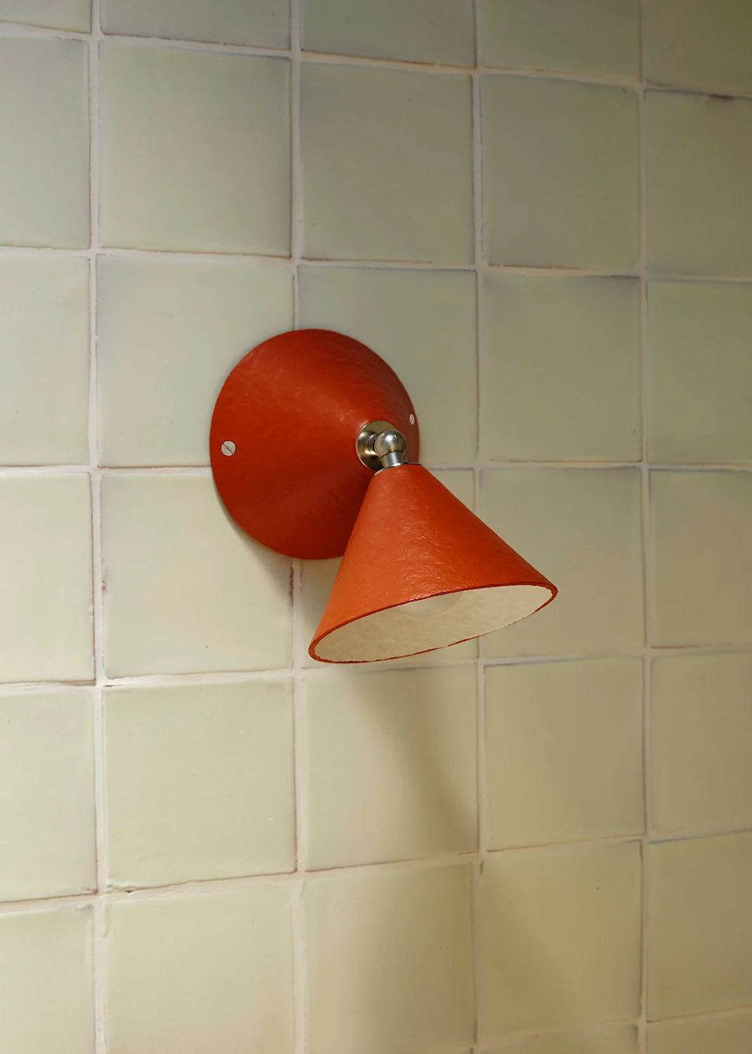A modern wall-mounted cone-shaped light fixture in terracotta orange, mounted on a beige tiled wall.