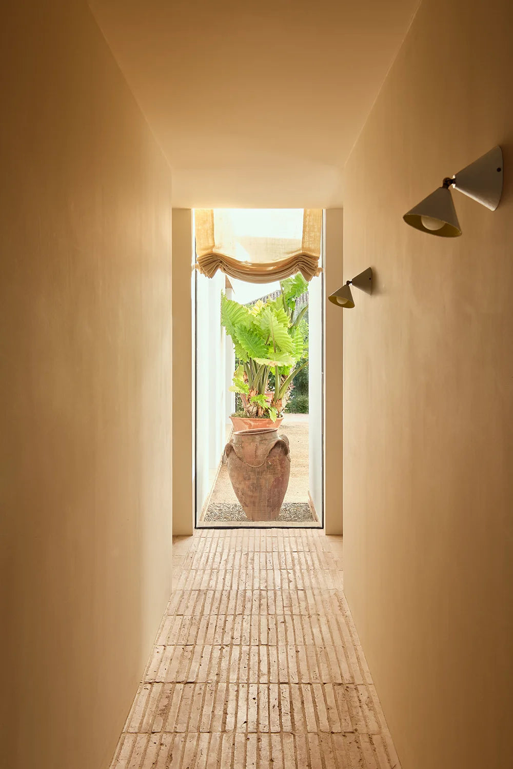 A long, narrow interior hallway with warm beige walls and a light tiled floor, leading to an open doorway where a large terracotta planter with green tropical foliage sits outdoors. Two white sconces line the right side of the hallway.