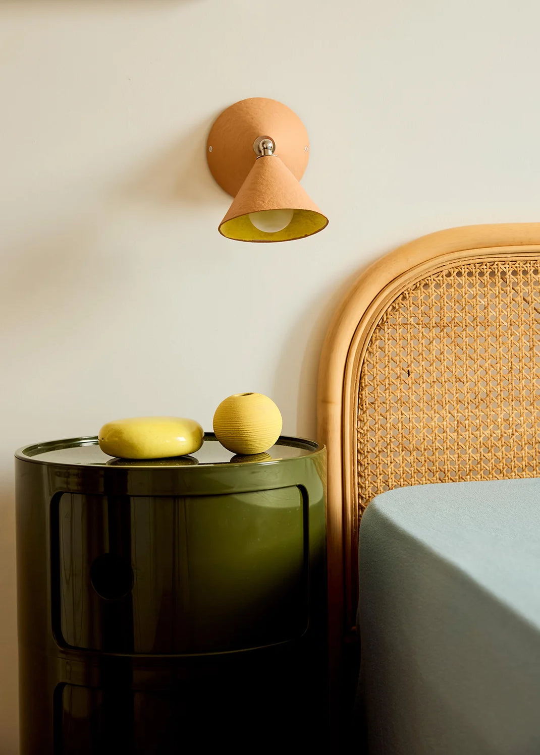 A peach-coloured wall light with a vibrant yellow interior hangs above a green side table and wicker bed.