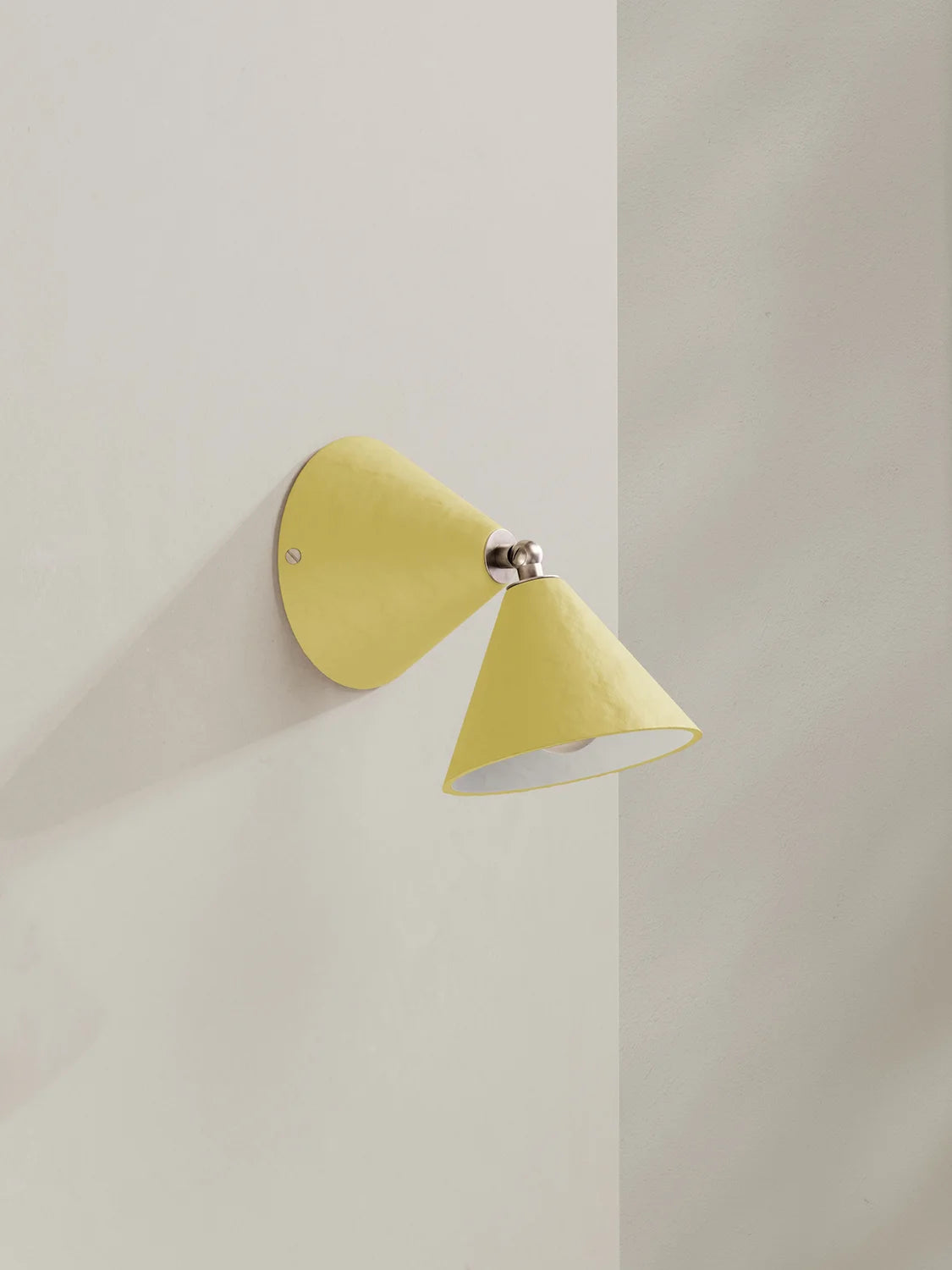 A modern wall-mounted sconce with a muted yellow cone-shaped shade and a brushed silver metal joint, shown against a minimalist off-white wall.