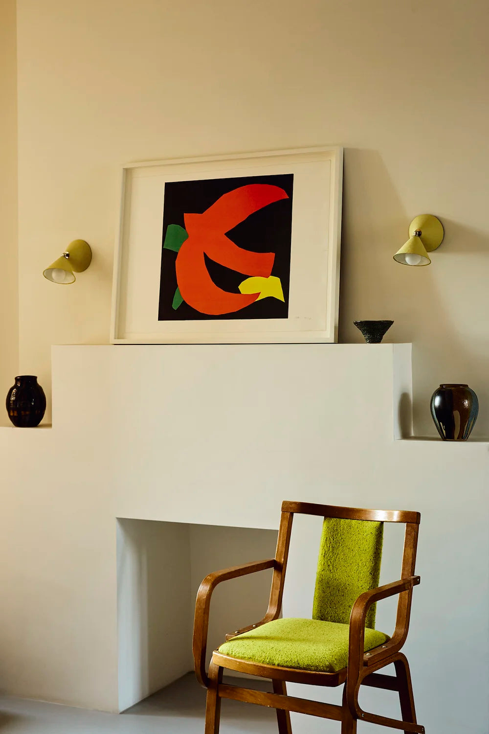 Modern interior with a white fireplace, abstract artwork, and a green cushioned chair. Two yellow wall sconces hang either side of the framed artwork.