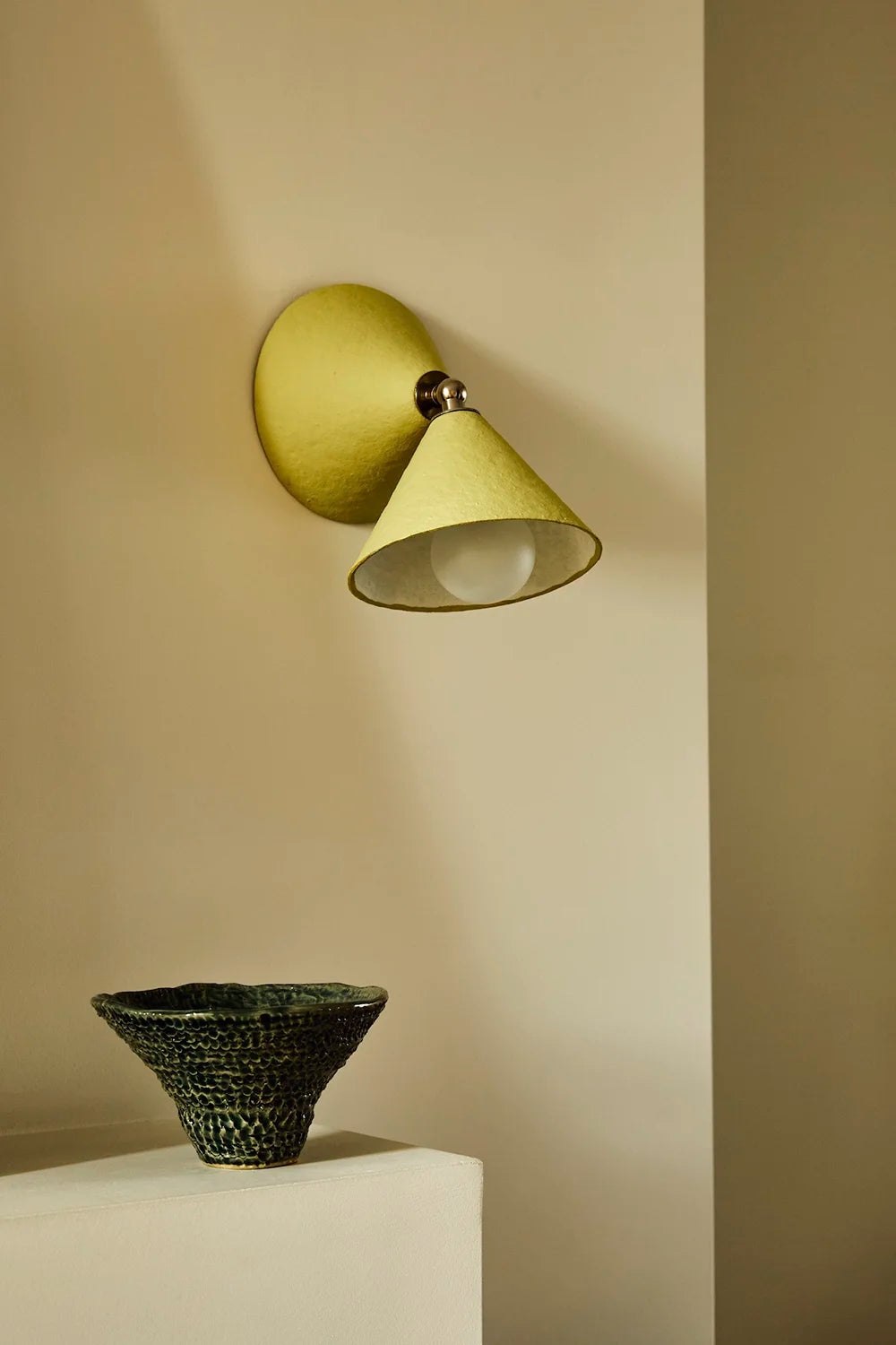 A modern wall sconce in a textured muted yellow, mounted on a beige wall above a white pedestal with a dark woven bowl.