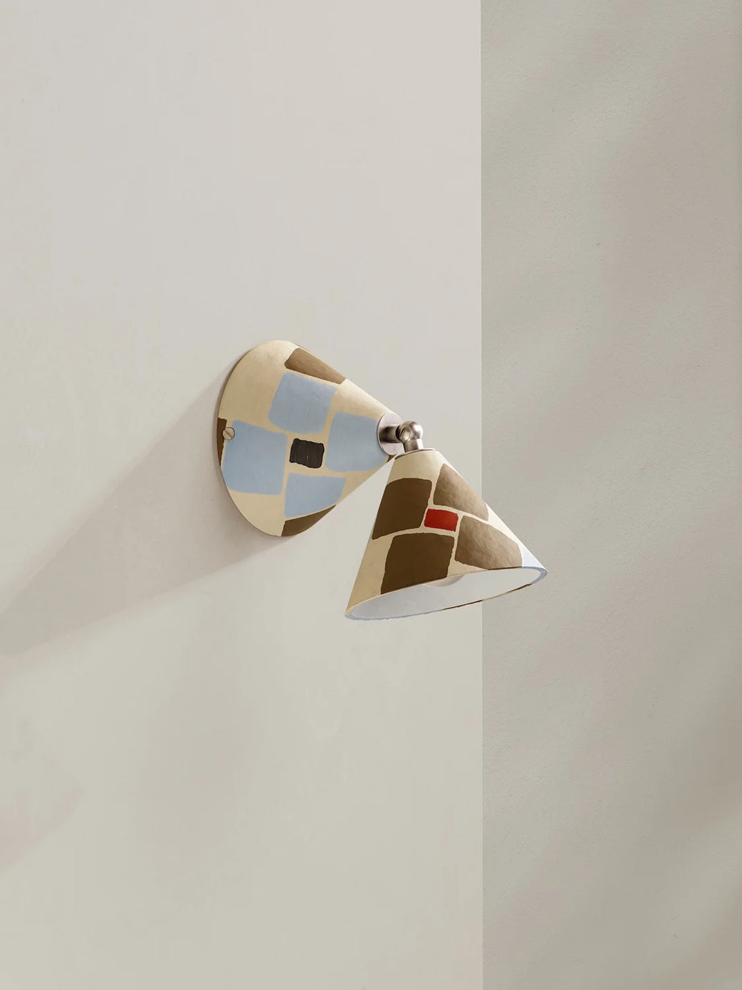 A modern wall-mounted sconce with a geometric mosaic pattern in beige, brown, light blue, and a small red accent, shown mounted on a neutral beige wall.