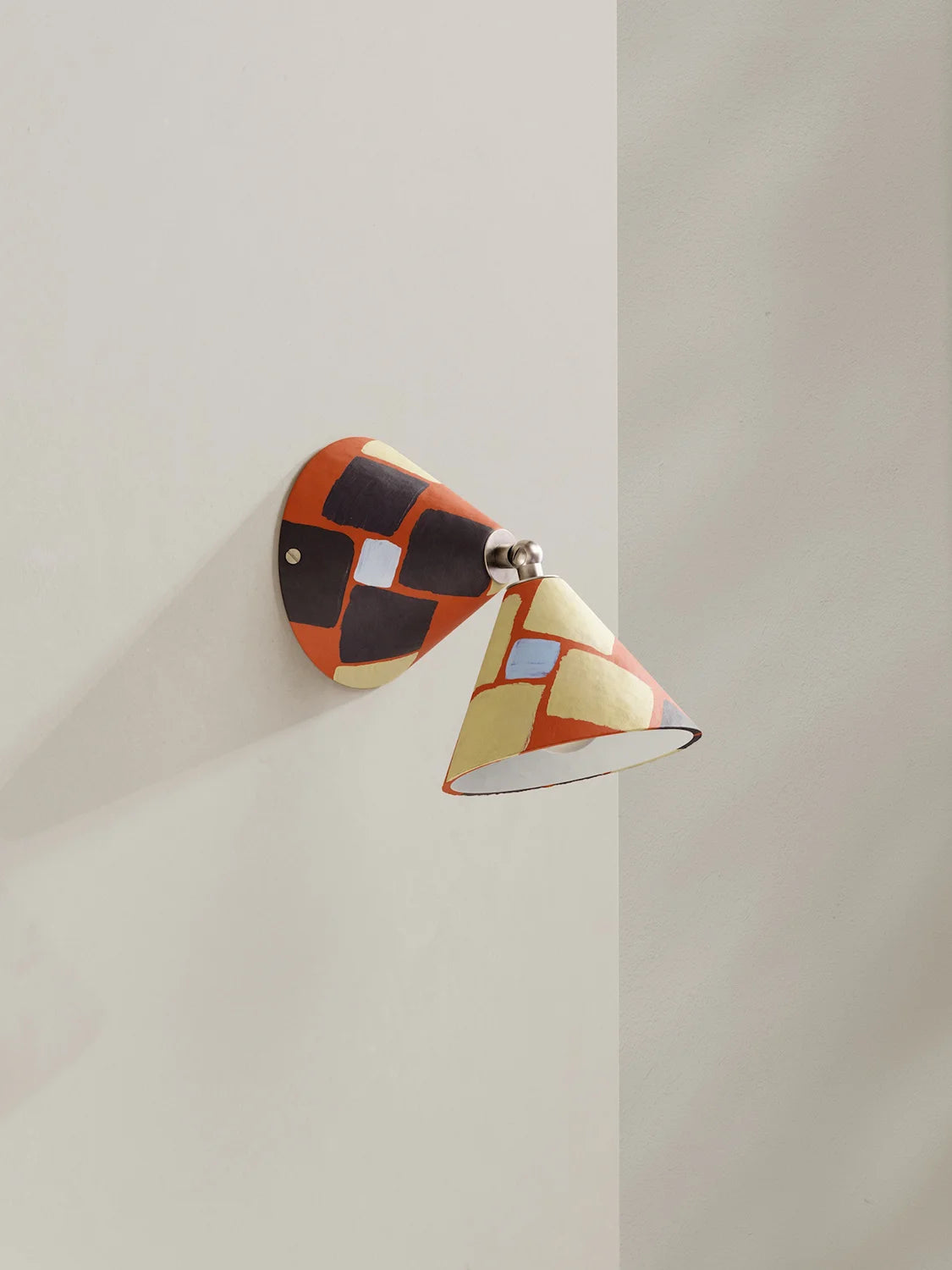 A modern wall-mounted sconce with a cone-shaped shade and a matching wall plate, decorated with an abstract geometric pattern of orange, black, cream, and light blue blocks, mounted on a neutral beige wall.