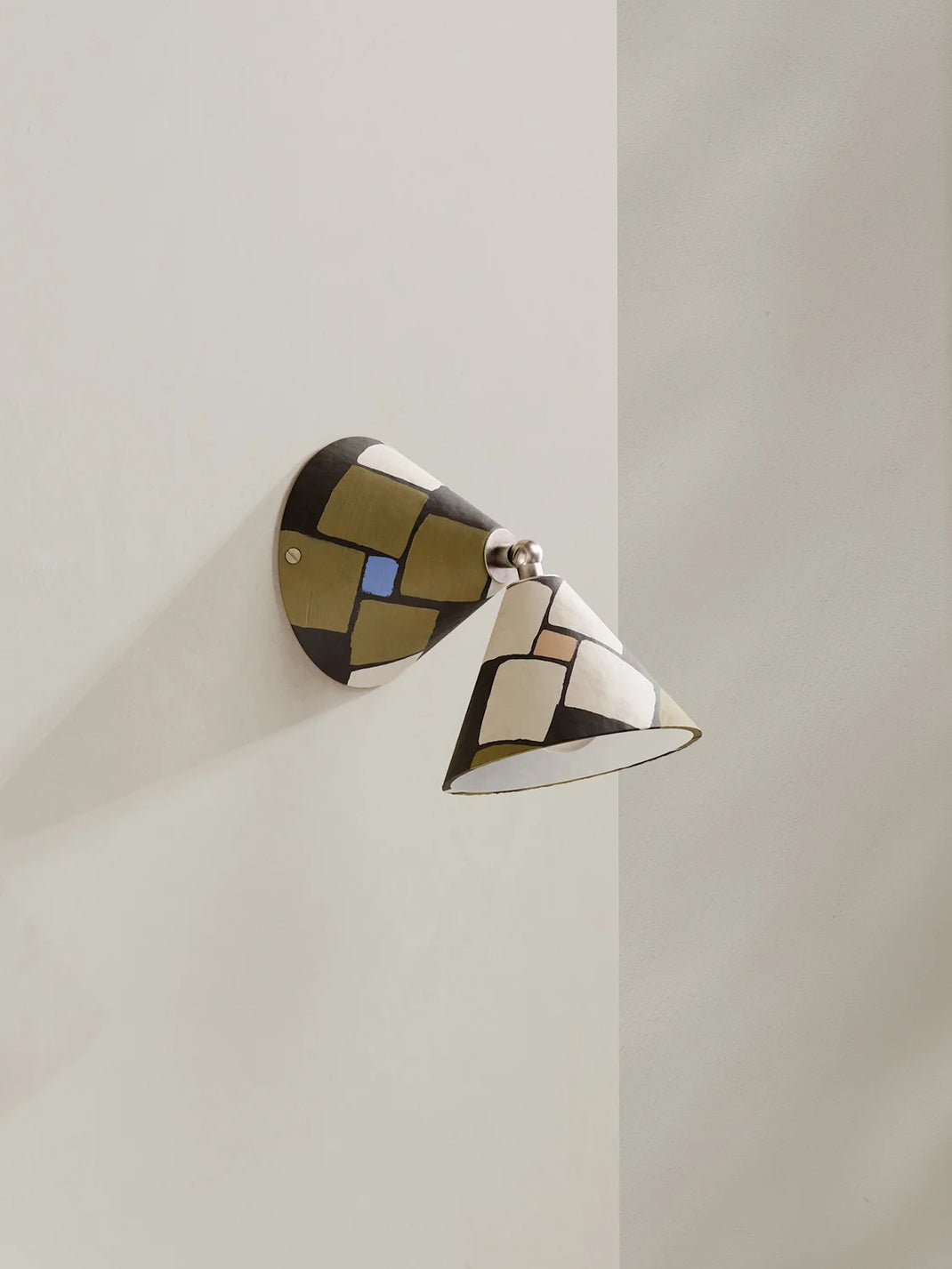 A modern geometric wall sconce with a conical shade and matching wall mount, decorated with a mosaic pattern of black, white, olive green, beige, and a small blue accent, mounted on a light beige wall.