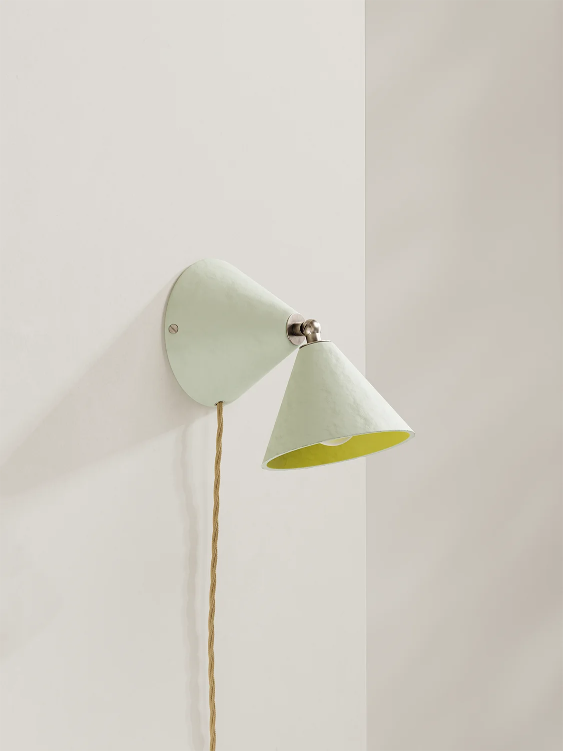 A modern wall sconce with an off-white outer shade and a bright yellow inner shade, mounted on a warm off-white wall with a visible cord.