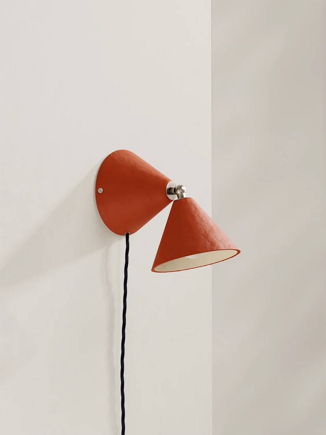 A modern cone-shaped wall sconce in terracotta orange with a dark blue cord, mounted on a clean white wall.