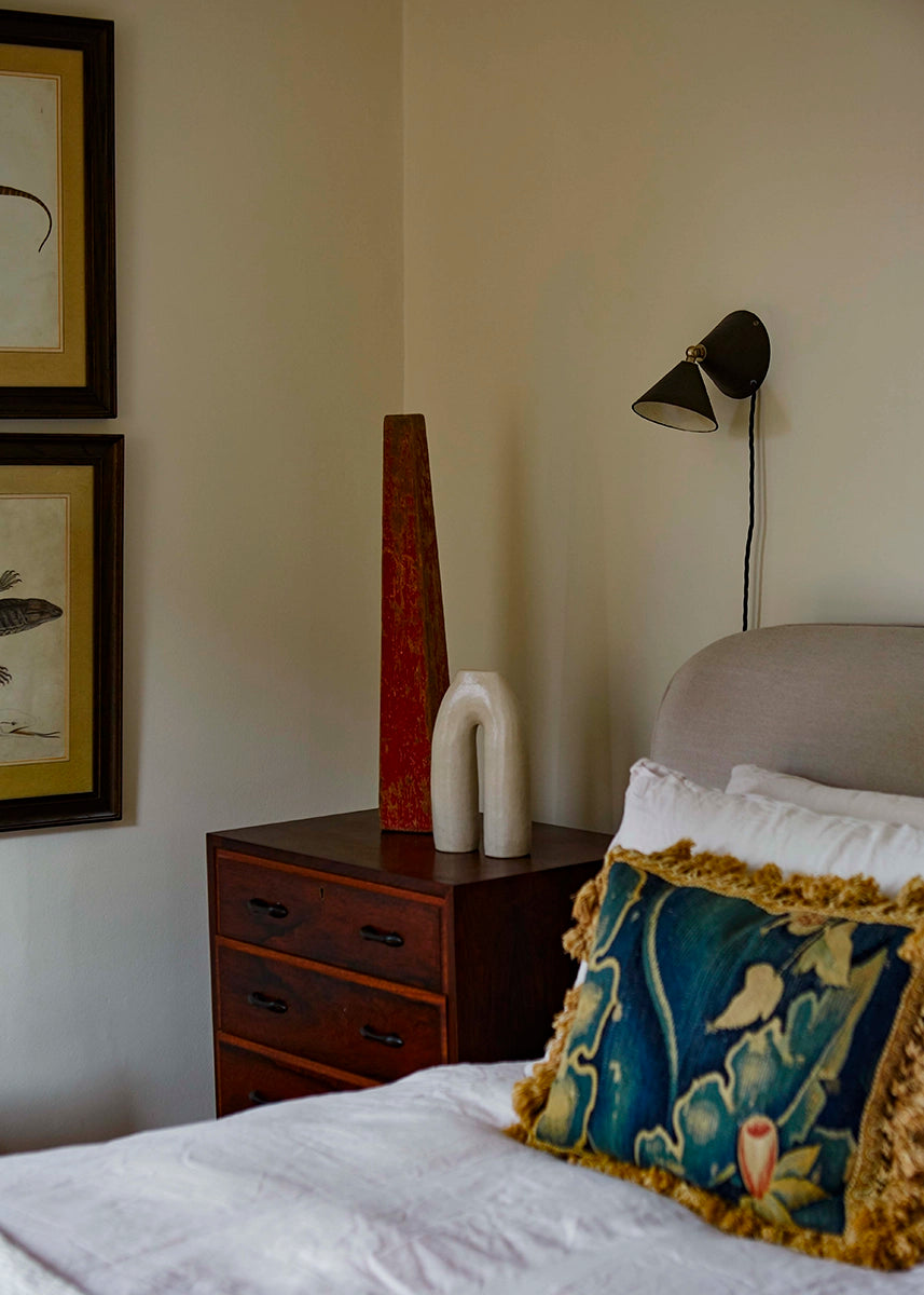A bedroom with decorative pillow, side table with ceramics, and a dark brown wall lamp