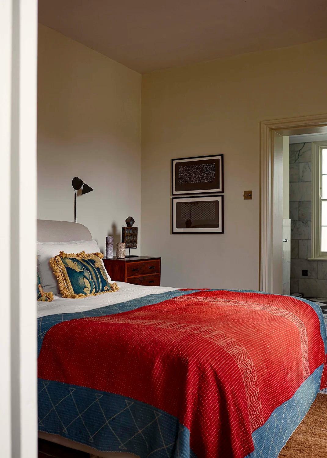 Bedroom with a red and blue blanket on a bed, framed pictures on the wall, and a dark brown wall lamp.