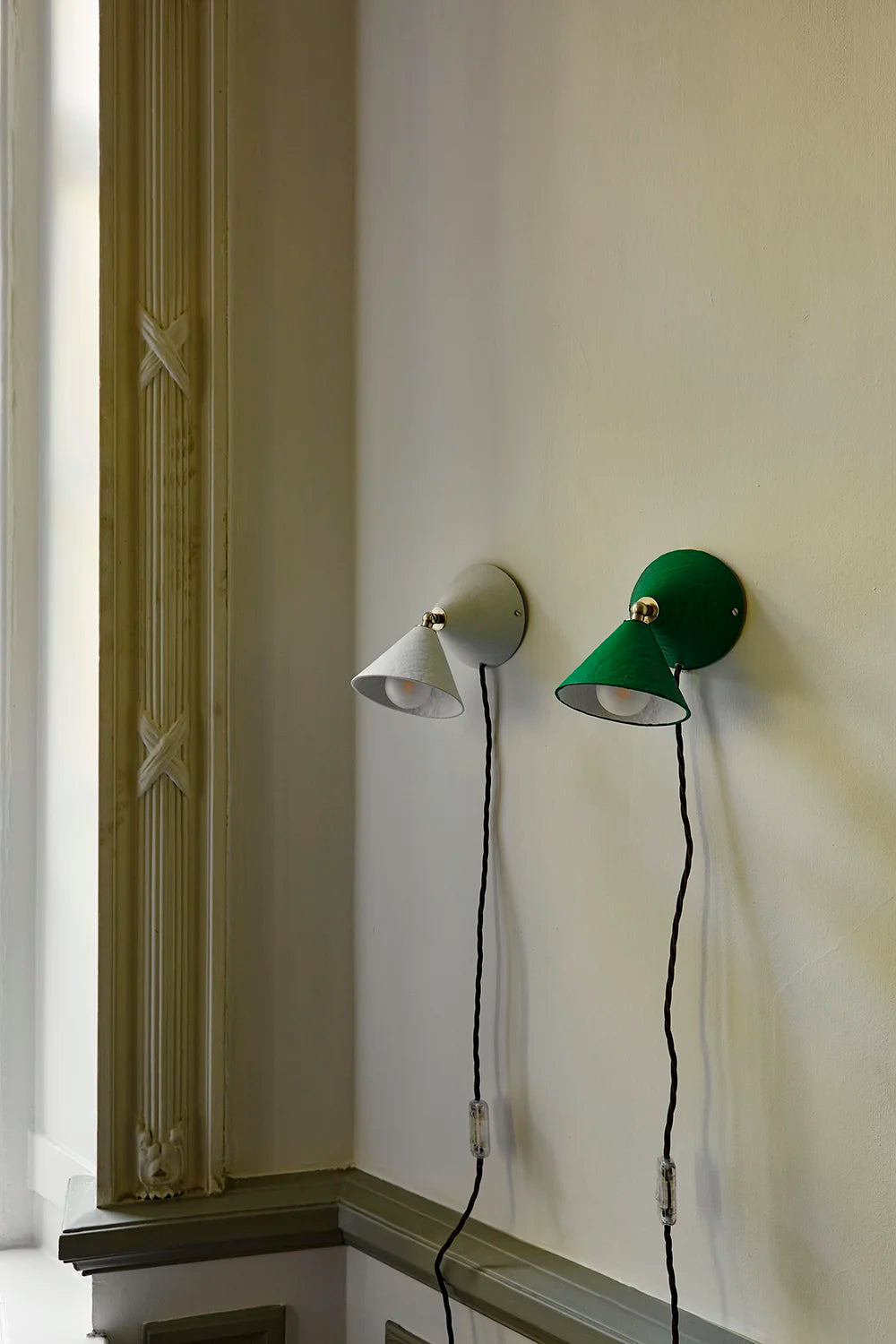 Two wall sconces mounted on a cream-colored wall, one in white and one in green, both with brown cords, positioned near a window frame.